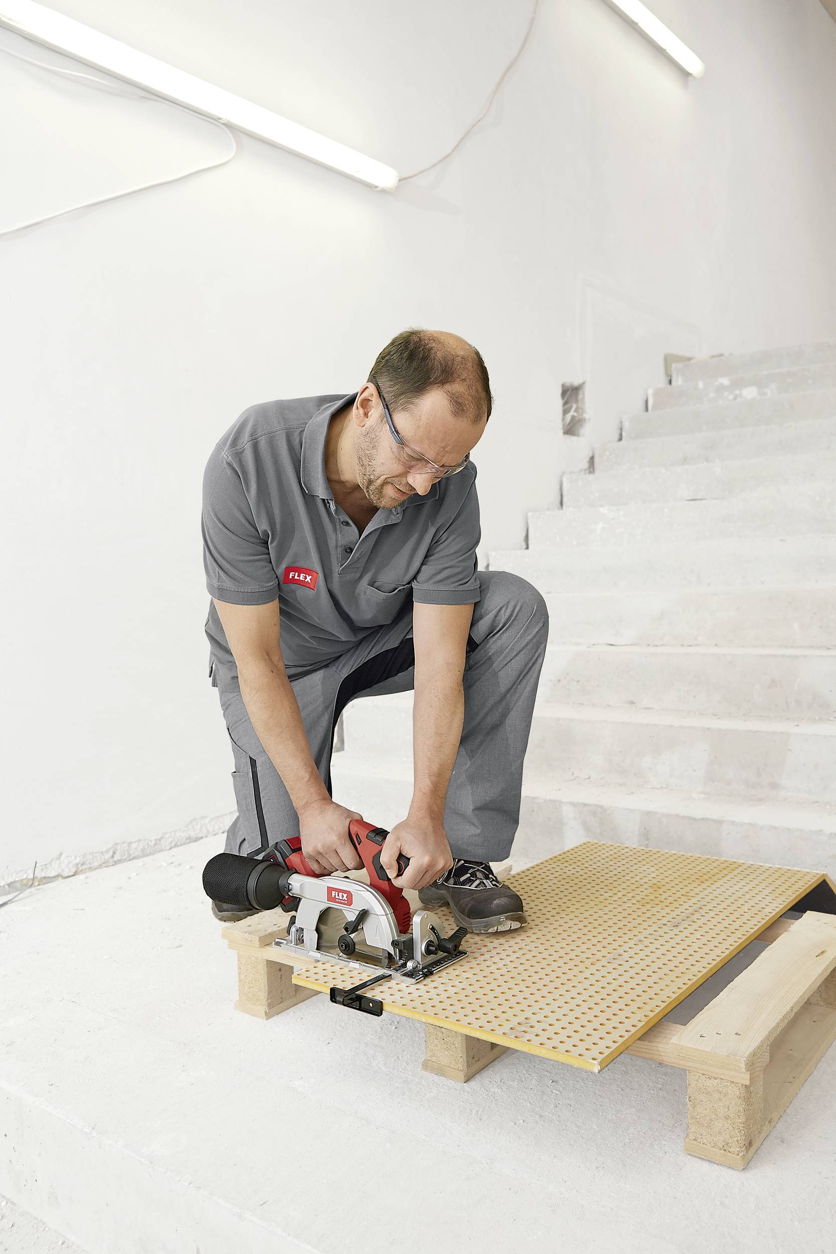 Un homme en tenue de travail grise utilise une scie circulaire portative pour couper une planche de bois, tout en étant agenouillé sur un escabeau.