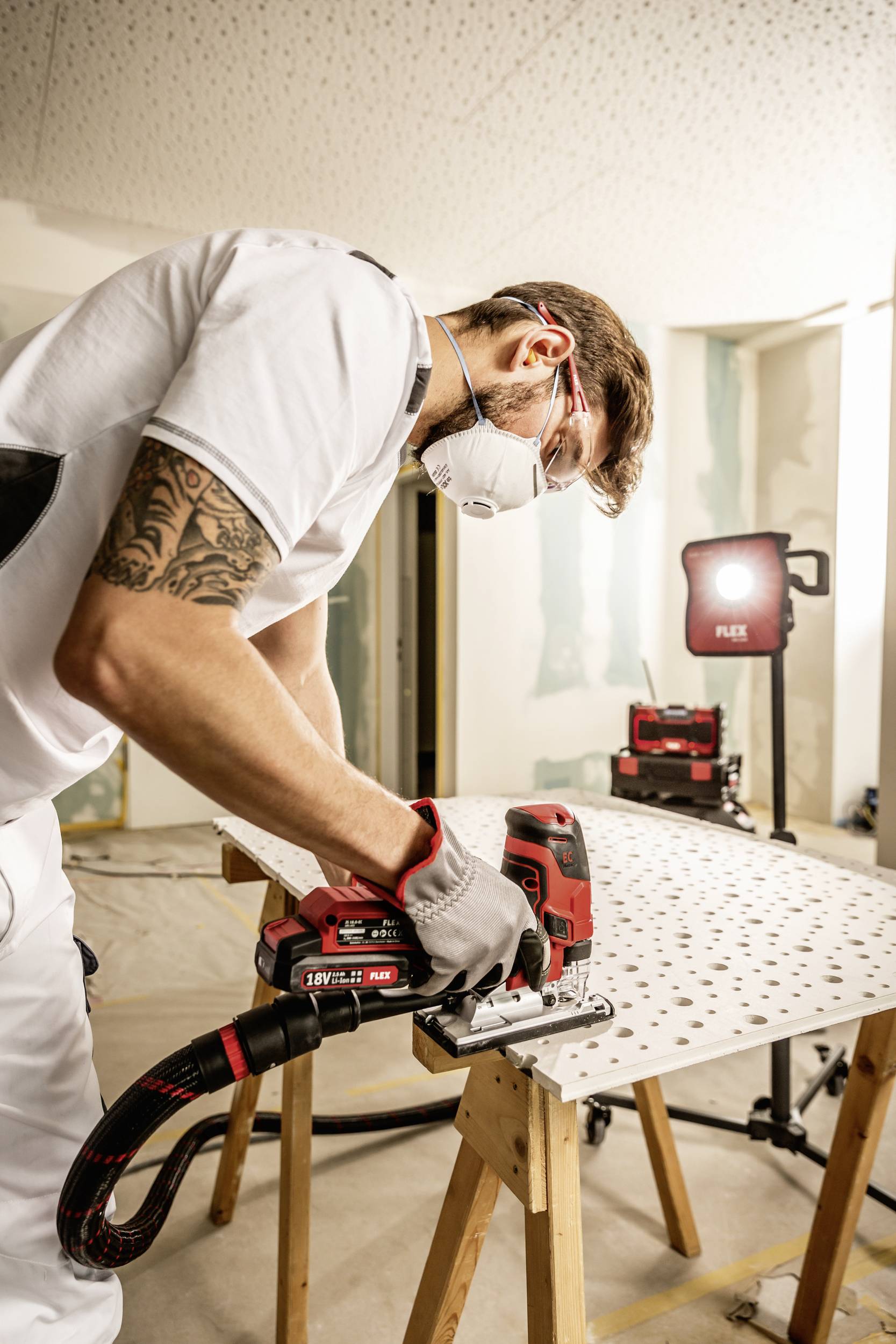 Un ouvrier portant un masque de protection et des outils travaille sur du matériel de chantier sur une table dans une pièce rénovée.