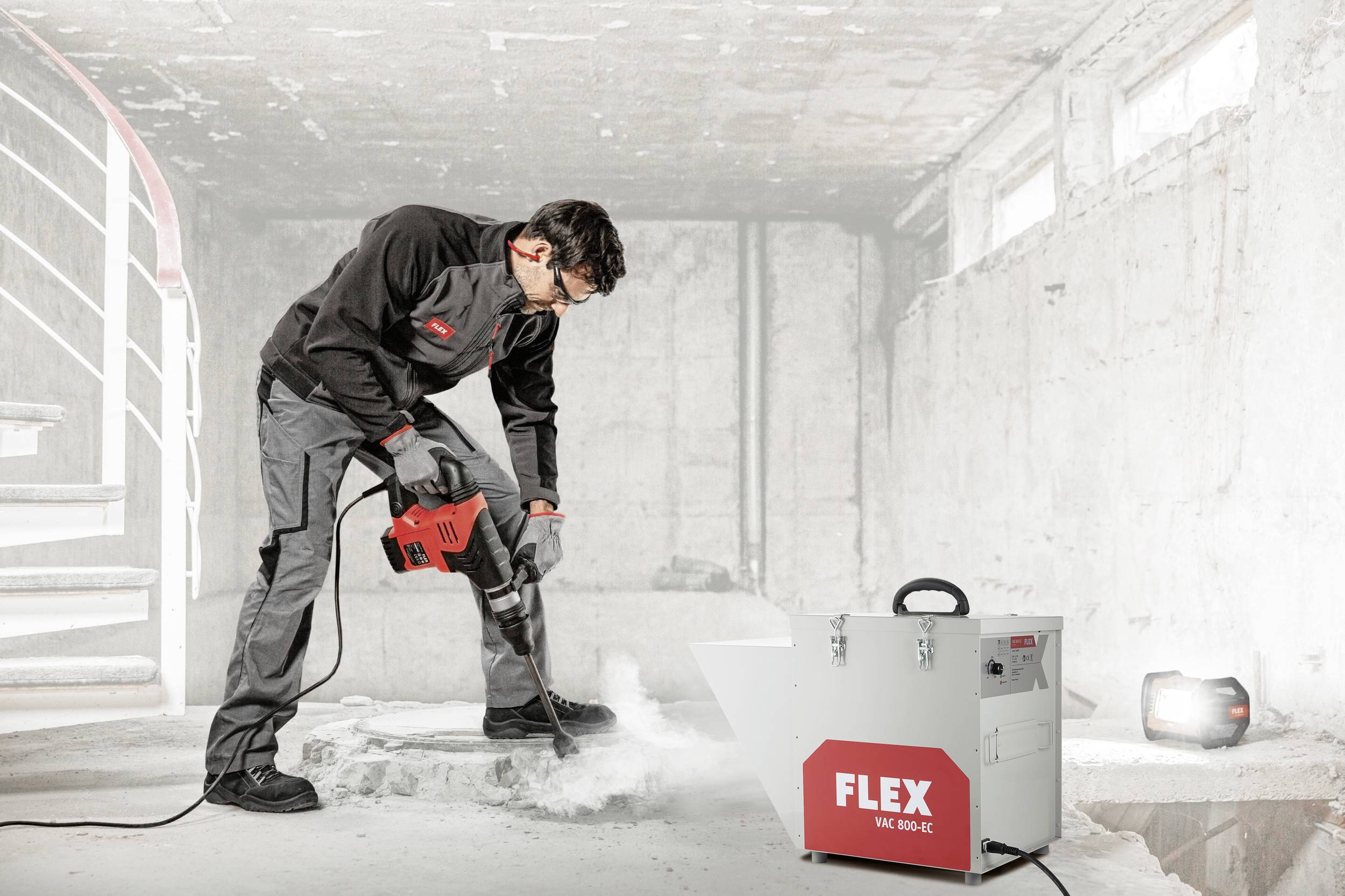 Un ouvrier en tenue de travail utilise un marteau-piqueur pour enlever du béton. Un aspirateur FLEX est posé à côté.