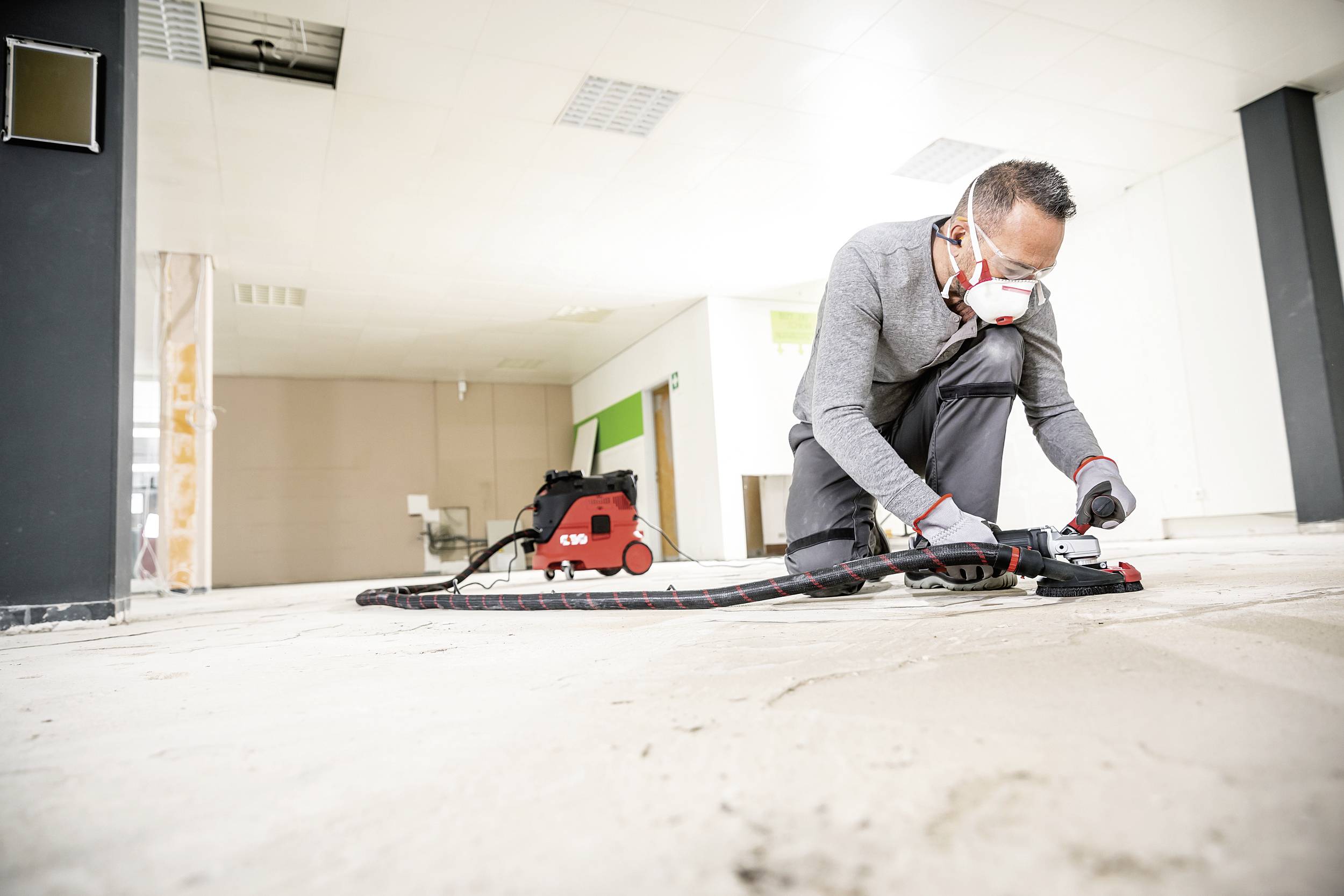 Un homme en tenue de protection est à genoux sur un sol poussiéreux et travaille avec un outil spécialisé. Des travaux de construction sont visibles en arrière-plan.