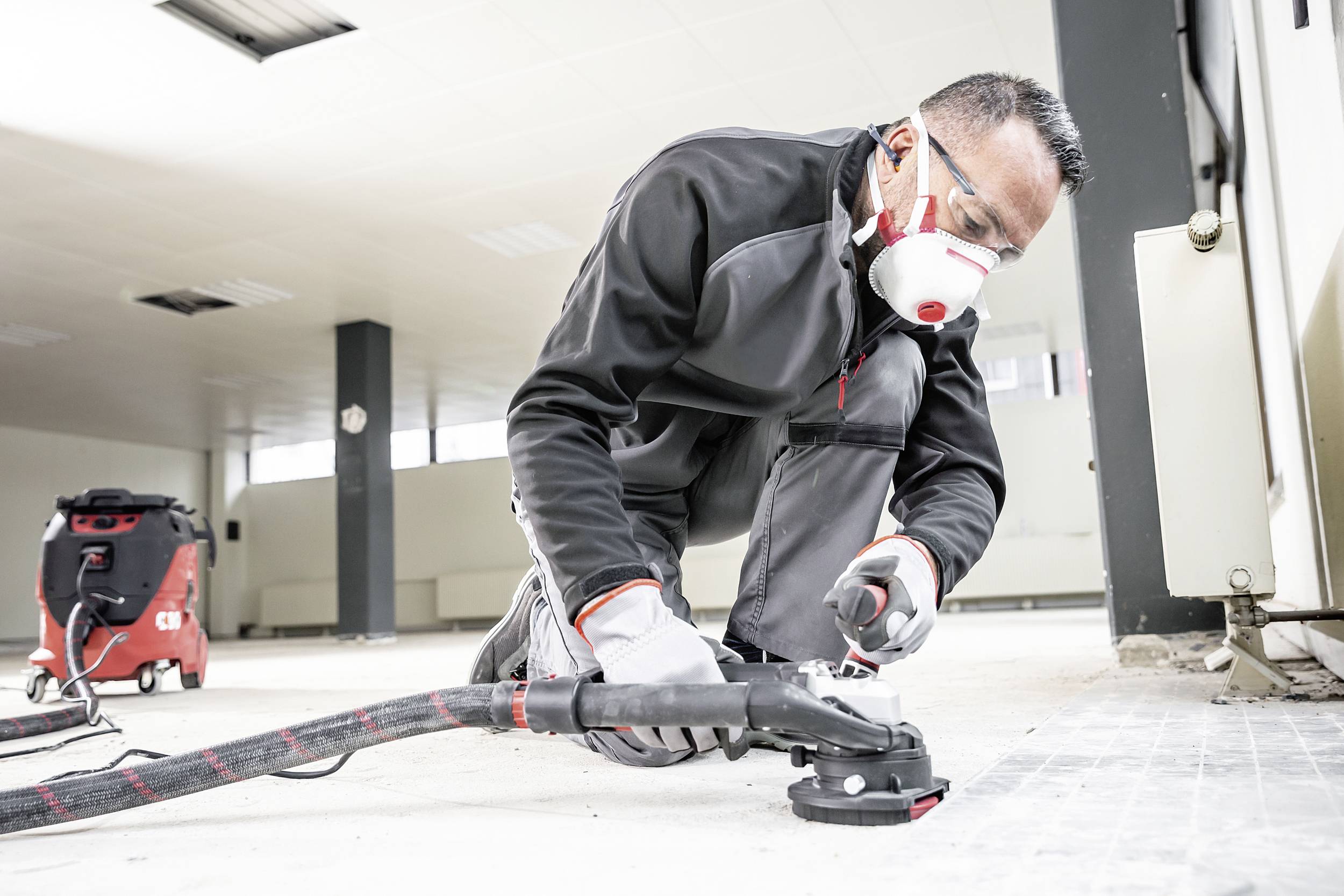 Un ouvrier porte des vêtements de protection et un masque alors qu'il travaille avec un outil de sol. Un aspirateur industriel est visible en arrière-plan.