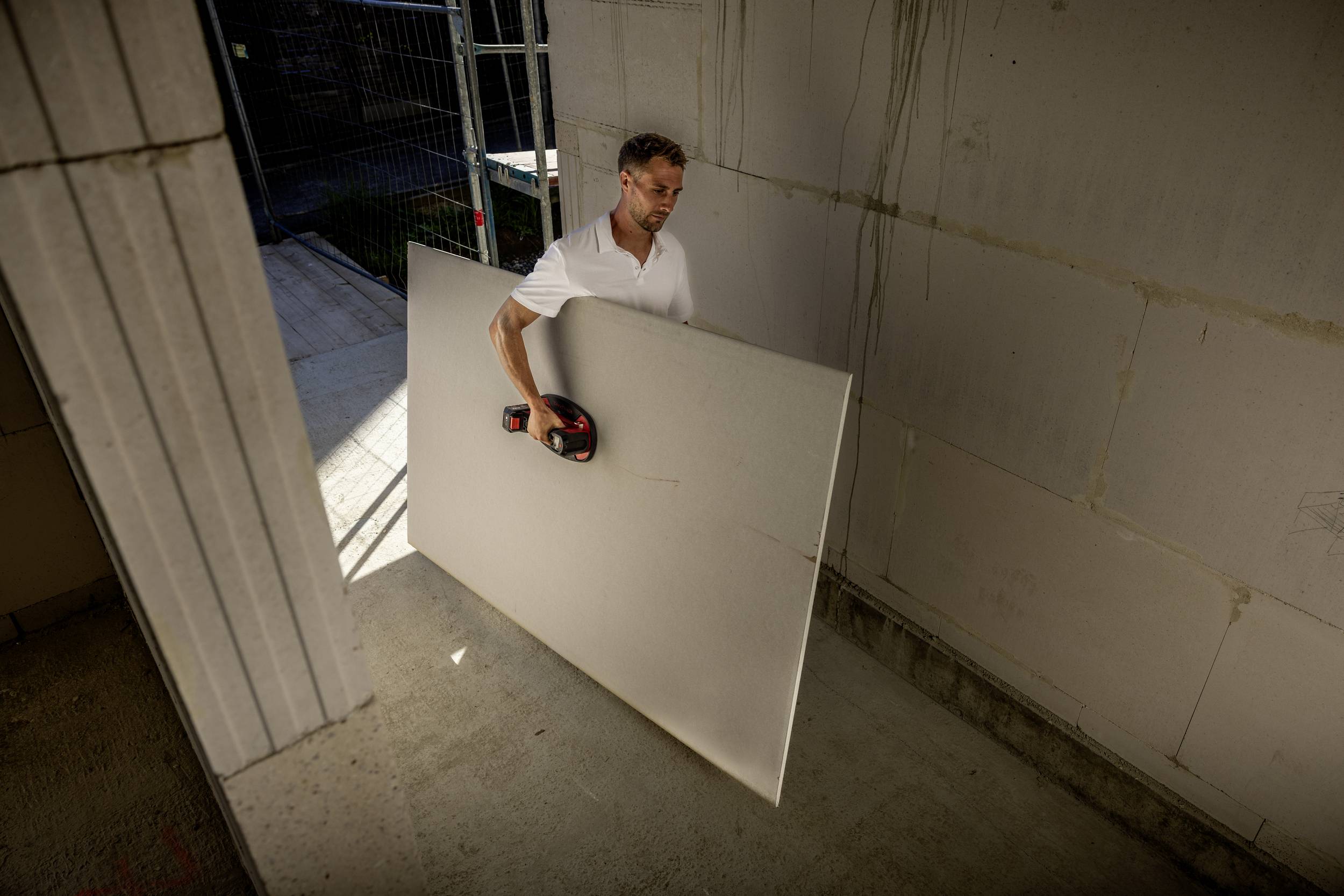 Une personne portant une chemise blanche soulève une grande plaque de plâtre dans une pièce inachevée, entourée de murs en cours de construction.