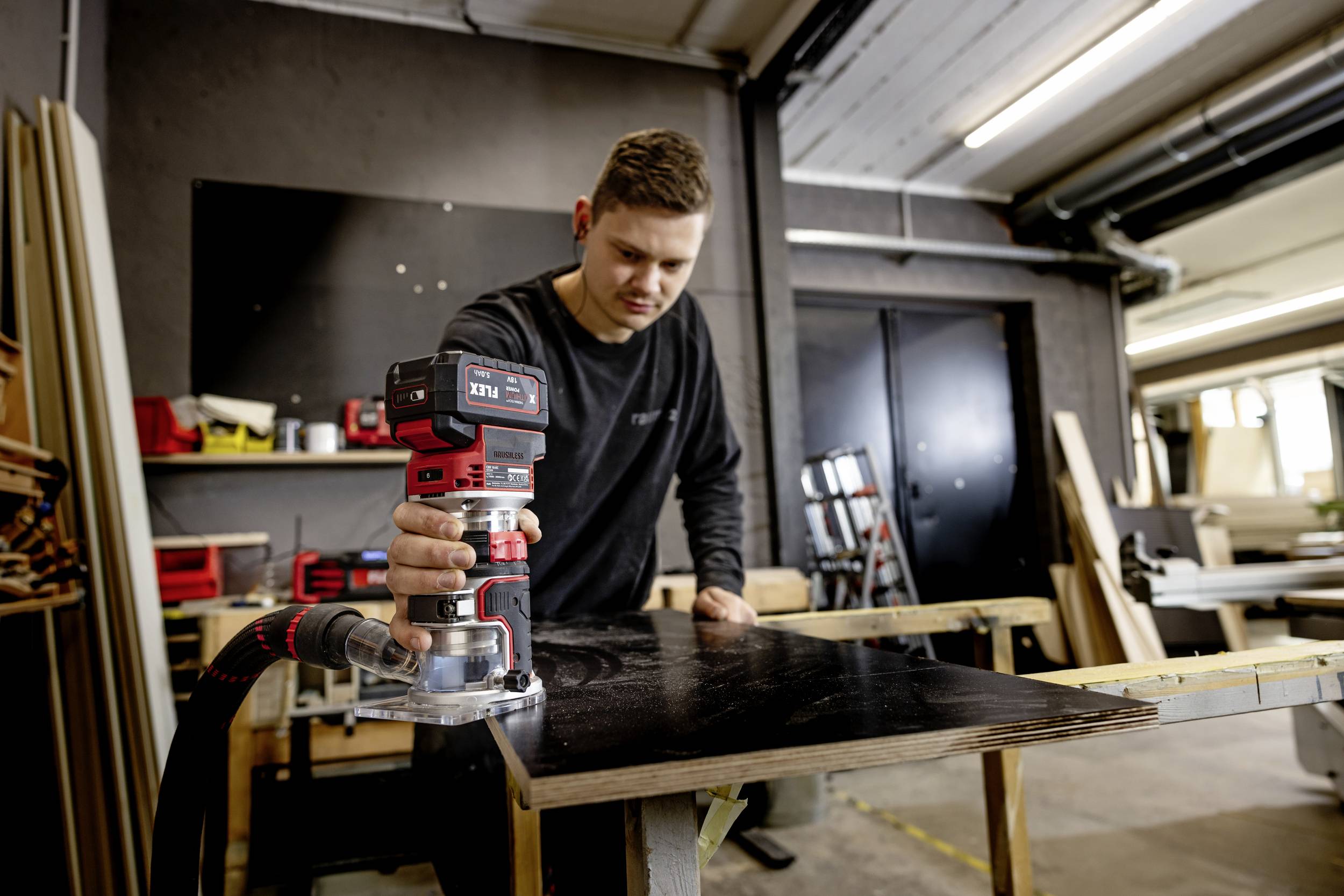 Un homme travaille une planche en bois sombre avec une ponceuse électrique dans un atelier. Des outils sont visibles en arrière-plan.