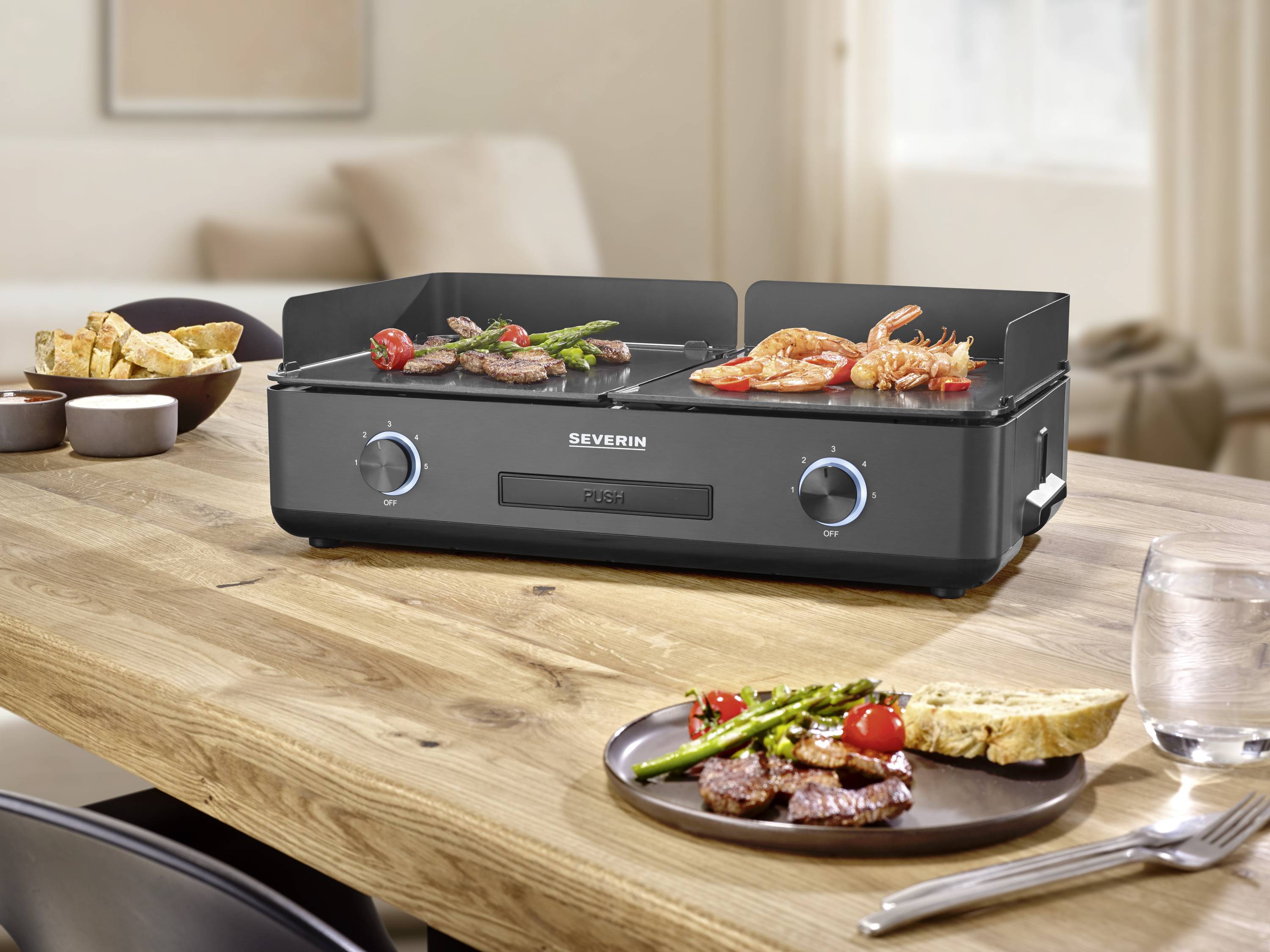 Un grill de table noir de Severin avec des légumes et de la viande grillés, entouré d'assiettes avec de la nourriture, est posé sur une table en bois dans une cuisine.