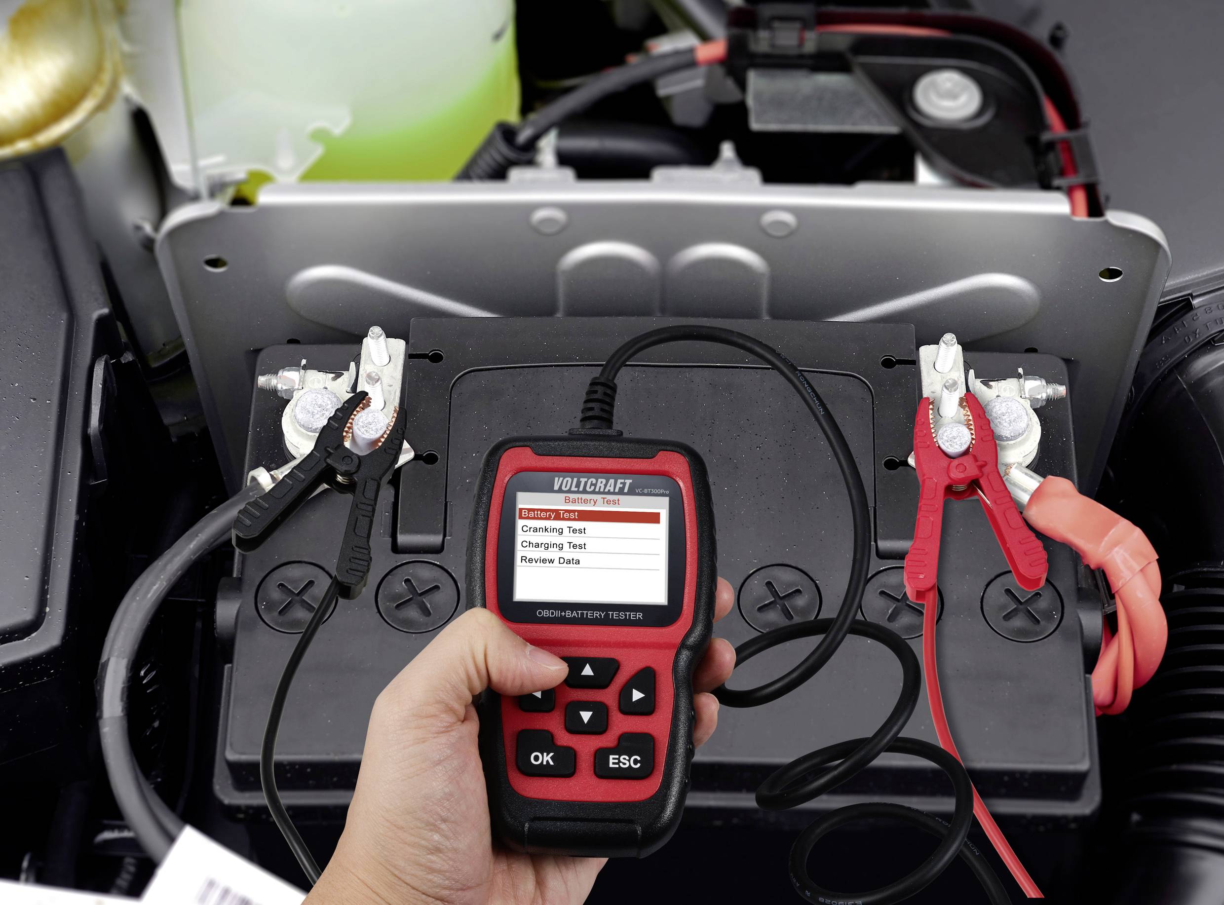 Une main tient un testeur de batterie connecté à une batterie de voiture. L'écran affiche différentes options de test.