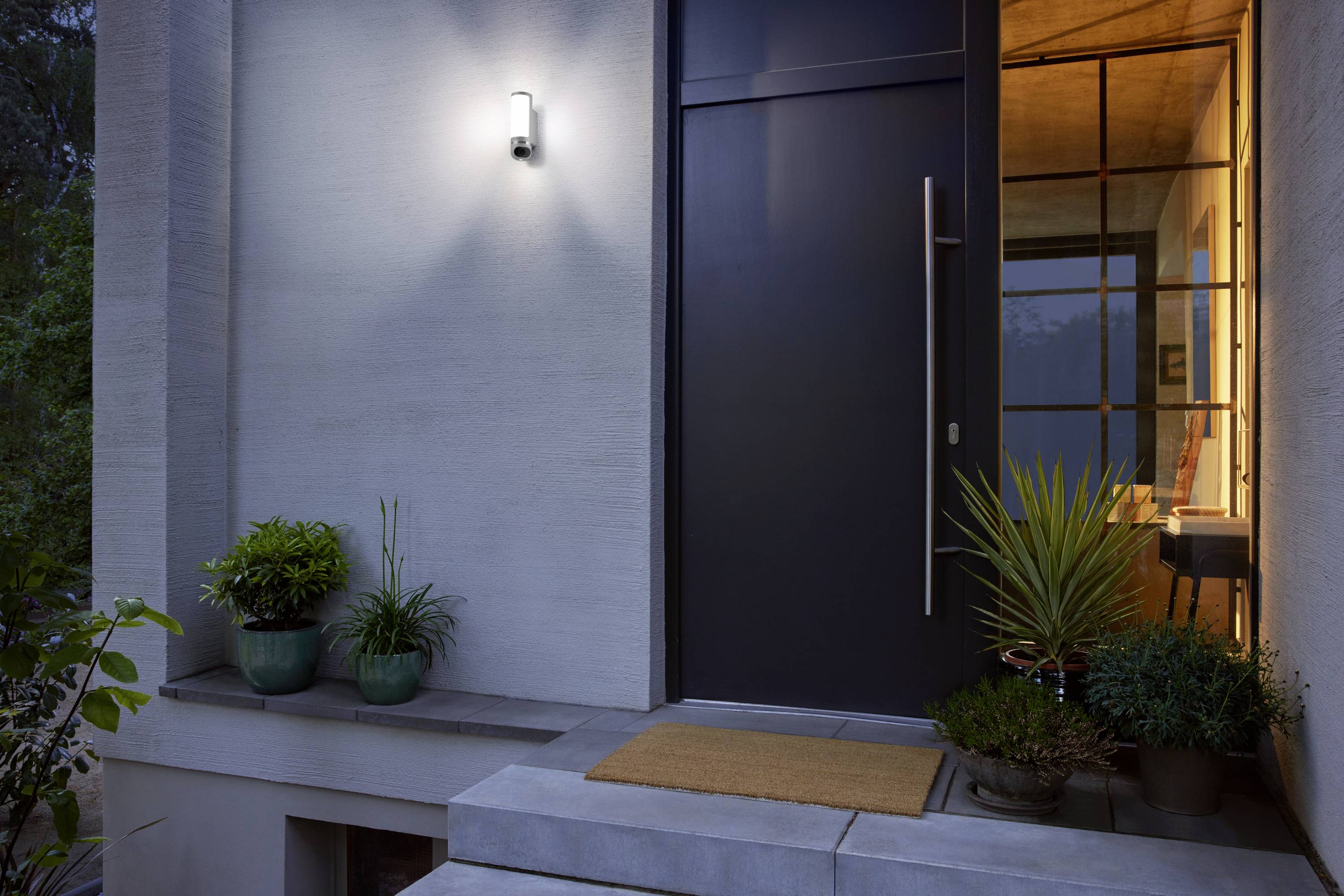 Une entrée de maison au design contemporain à la tombée du jour, éclairée par une applique murale, avec des plantes en pot et un tapis d'entrée marron.