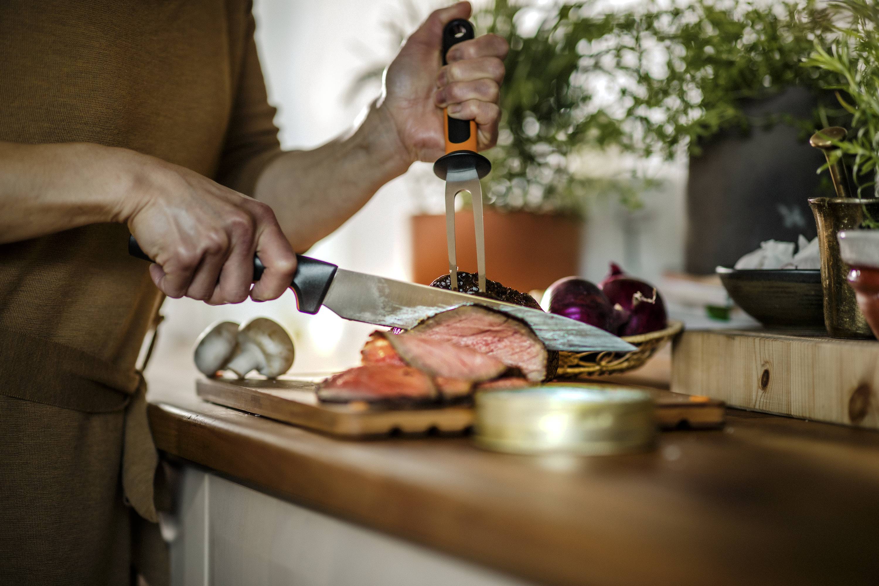 Une personne découpe un morceau de viande cuite sur une planche en bois dans une cuisine, entourée de légumes et d'herbes.