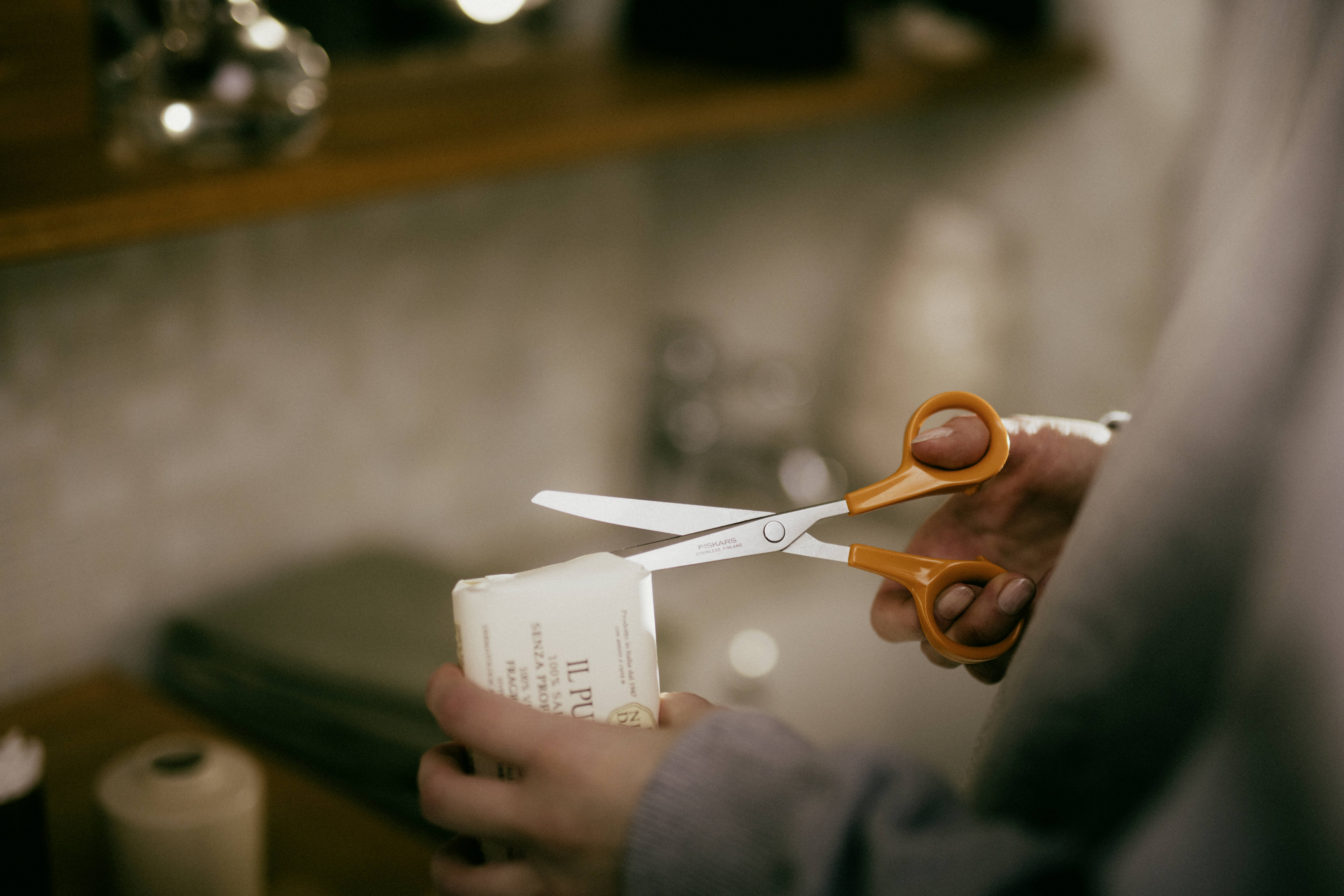 Une personne coupe l'emballage d'un savon avec des ciseaux. La scène se déroule dans une salle de bain avec un arrière-plan flou.