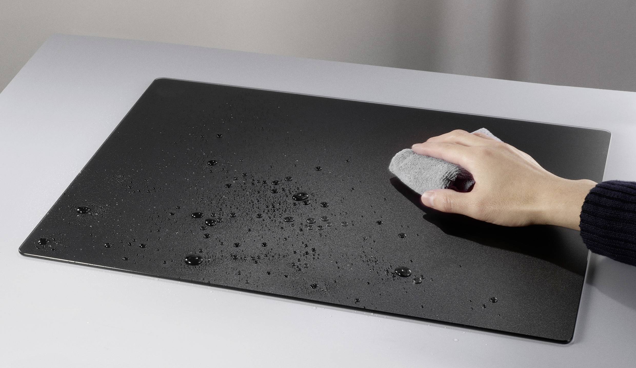 Une main essuie des gouttelettes d'eau sur une surface noire rectangulaire posée sur une table grise à l'aide d'un chiffon gris.