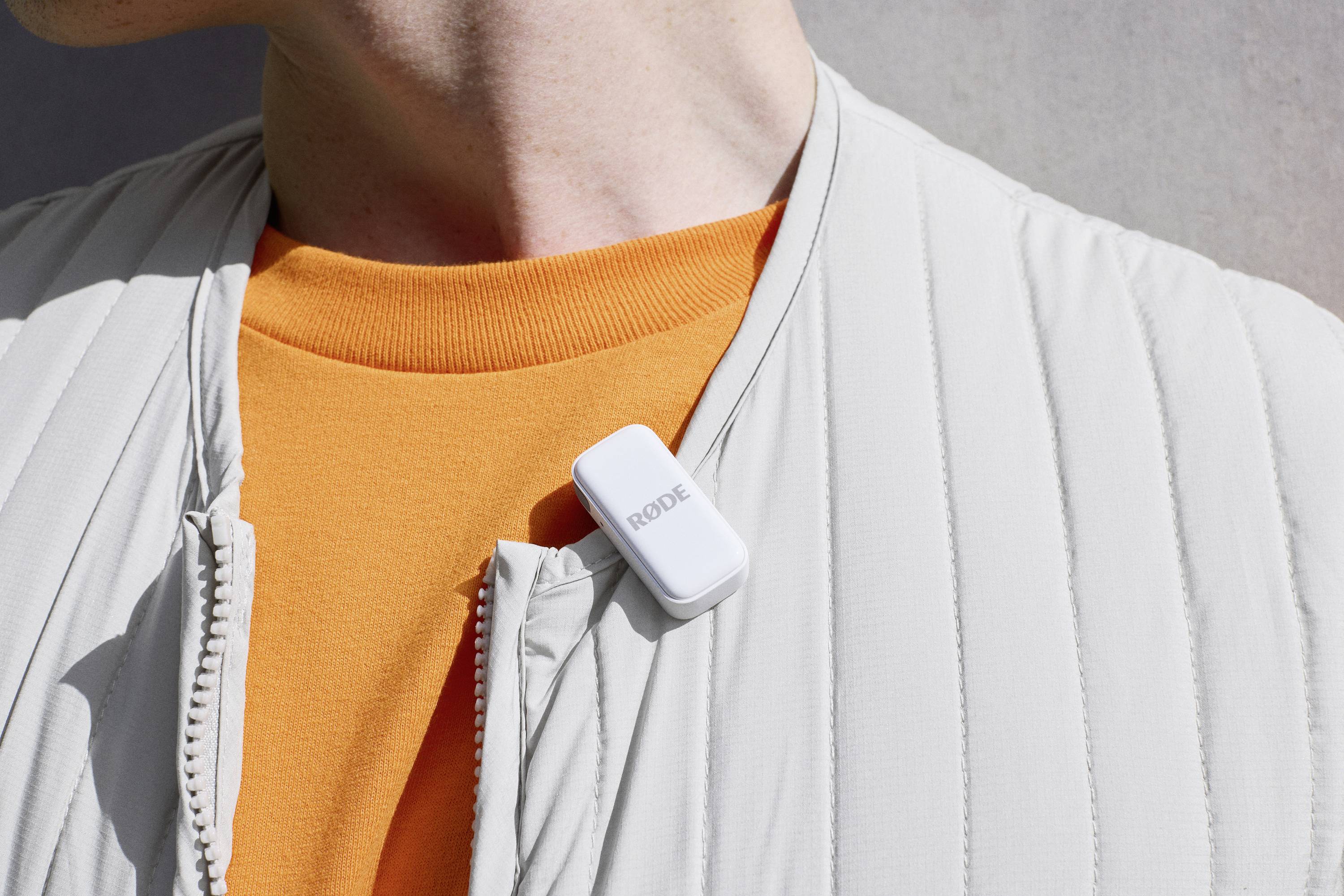Un microphone blanc sans fil est fixé à une veste gris clair devant un pull orange.