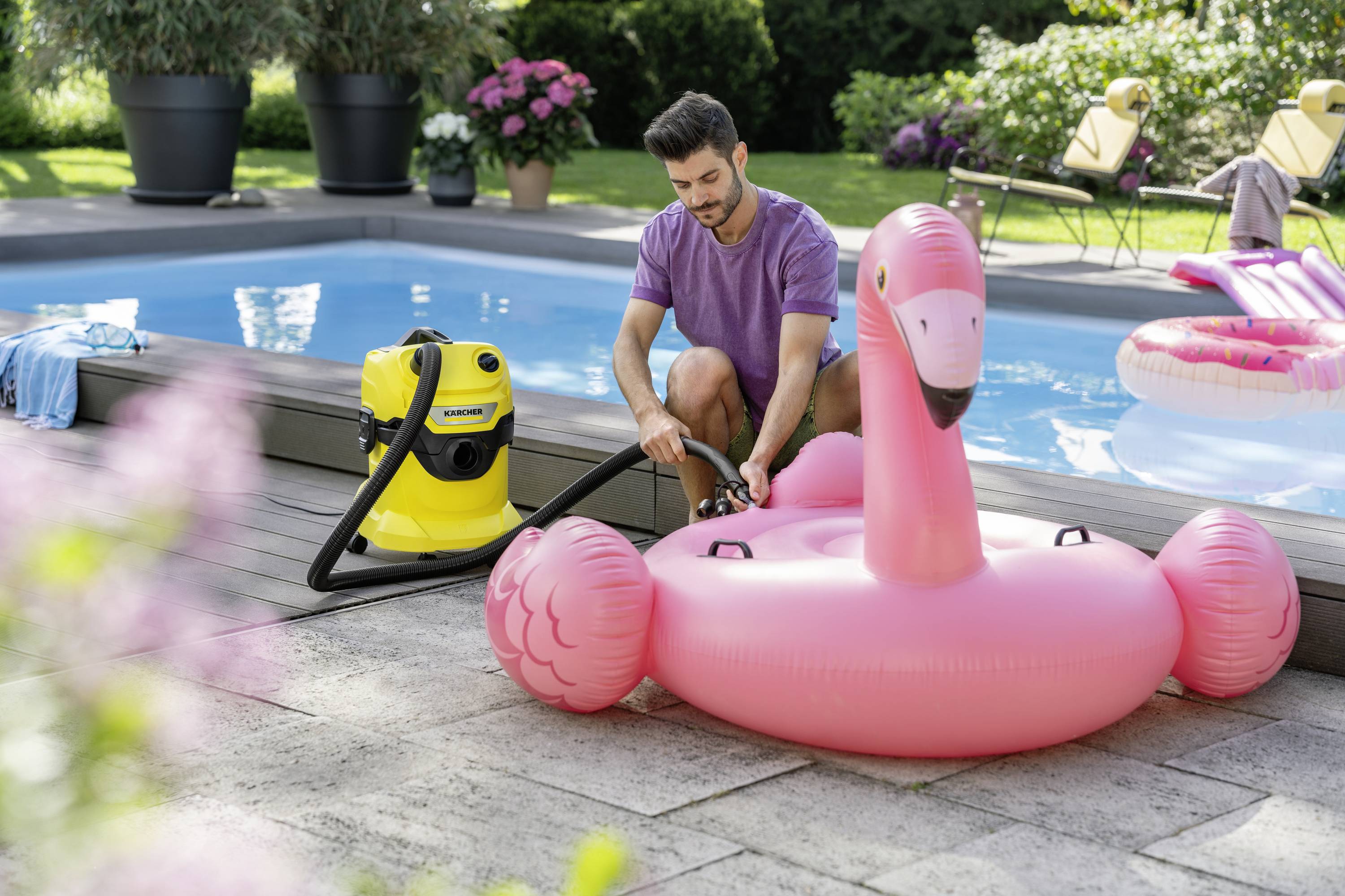 Un homme gonfle un grand flotteur de piscine en forme de flamant rose à l'aide d'un appareil jaune. Des plantes sont visibles en arrière-plan.
