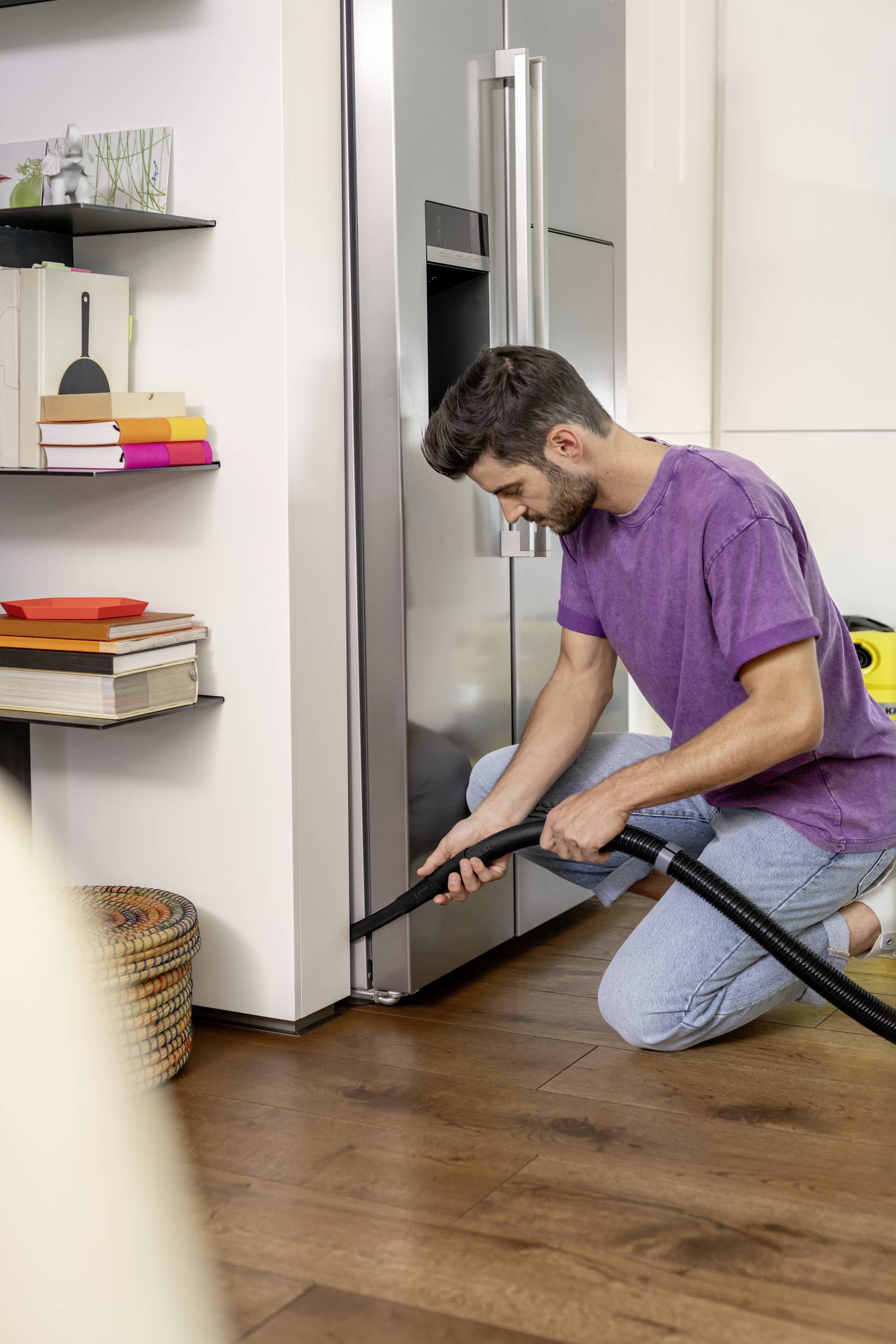Un homme est à genoux sur un plancher en bois et aspire le coin d'une cuisine moderne avec un aspirateur. Des étagères avec des livres sont visibles en arrière-plan.