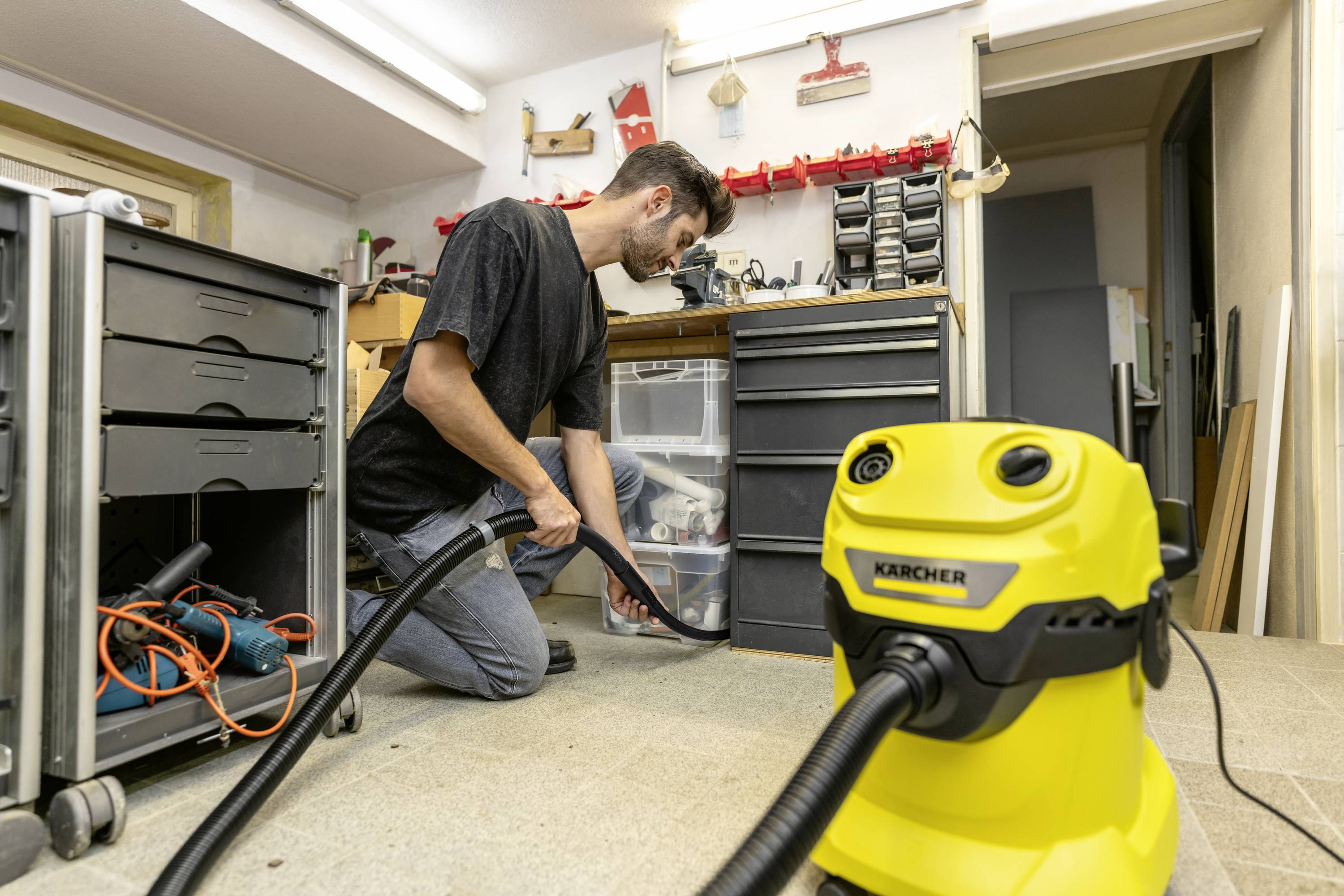 Un homme est agenouillé dans un atelier et utilise un aspirateur jaune pour nettoyer le sol. Des outils sont visibles en arrière-plan.