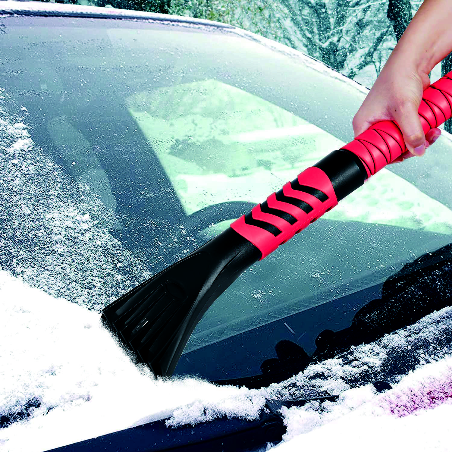 Une main utilise un grattoir à glace avec une poignée rouge pour enlever la neige et la glace du pare-brise d'une voiture.