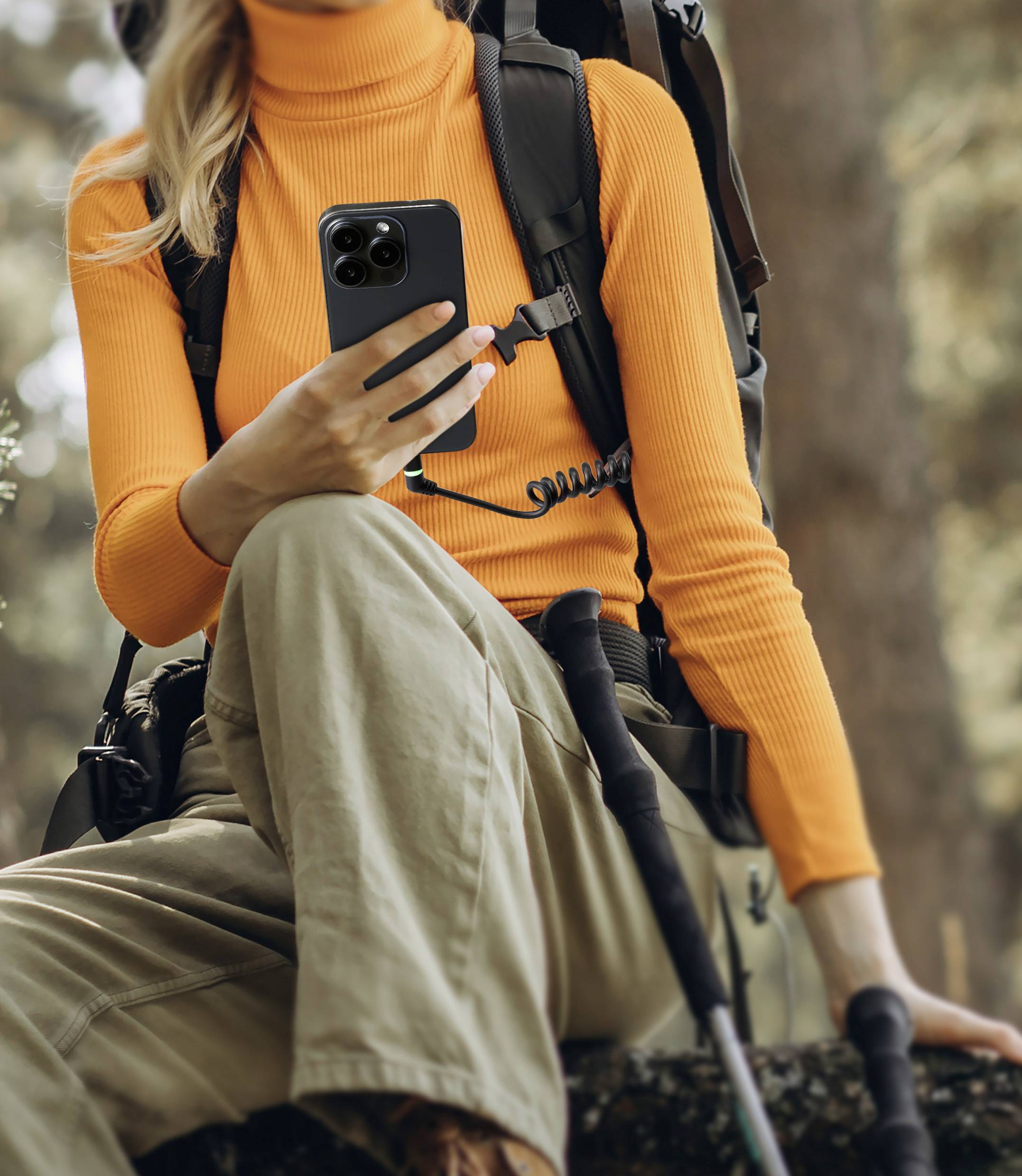 Une personne portant un pull orange est assise à l'extérieur, un sac à dos sur le dos, et regarde un smartphone.