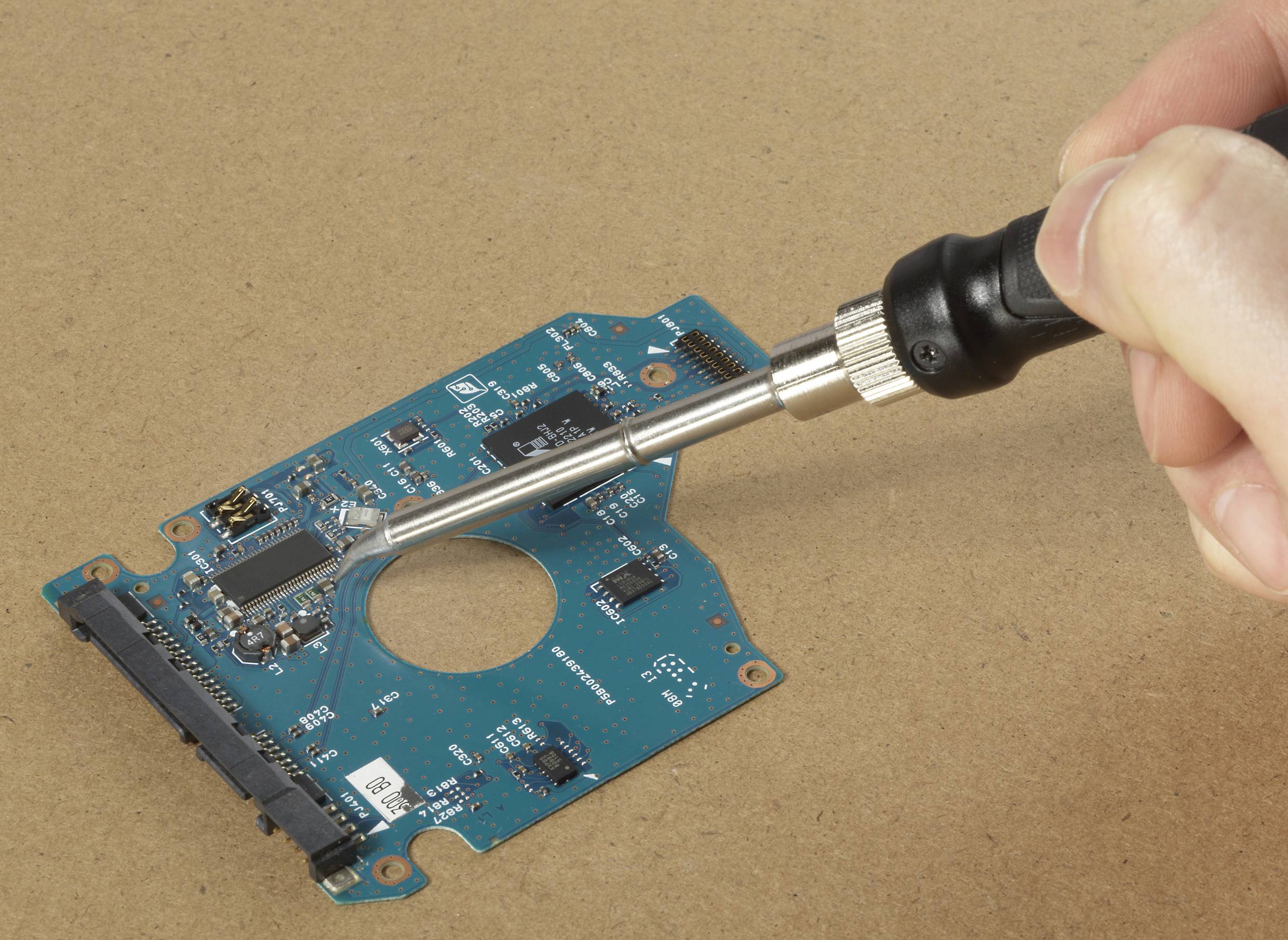 Une main soude des composants sur un circuit imprimé vert à l'aide d'un fer à souder. Le circuit imprimé contient plusieurs composants électroniques.