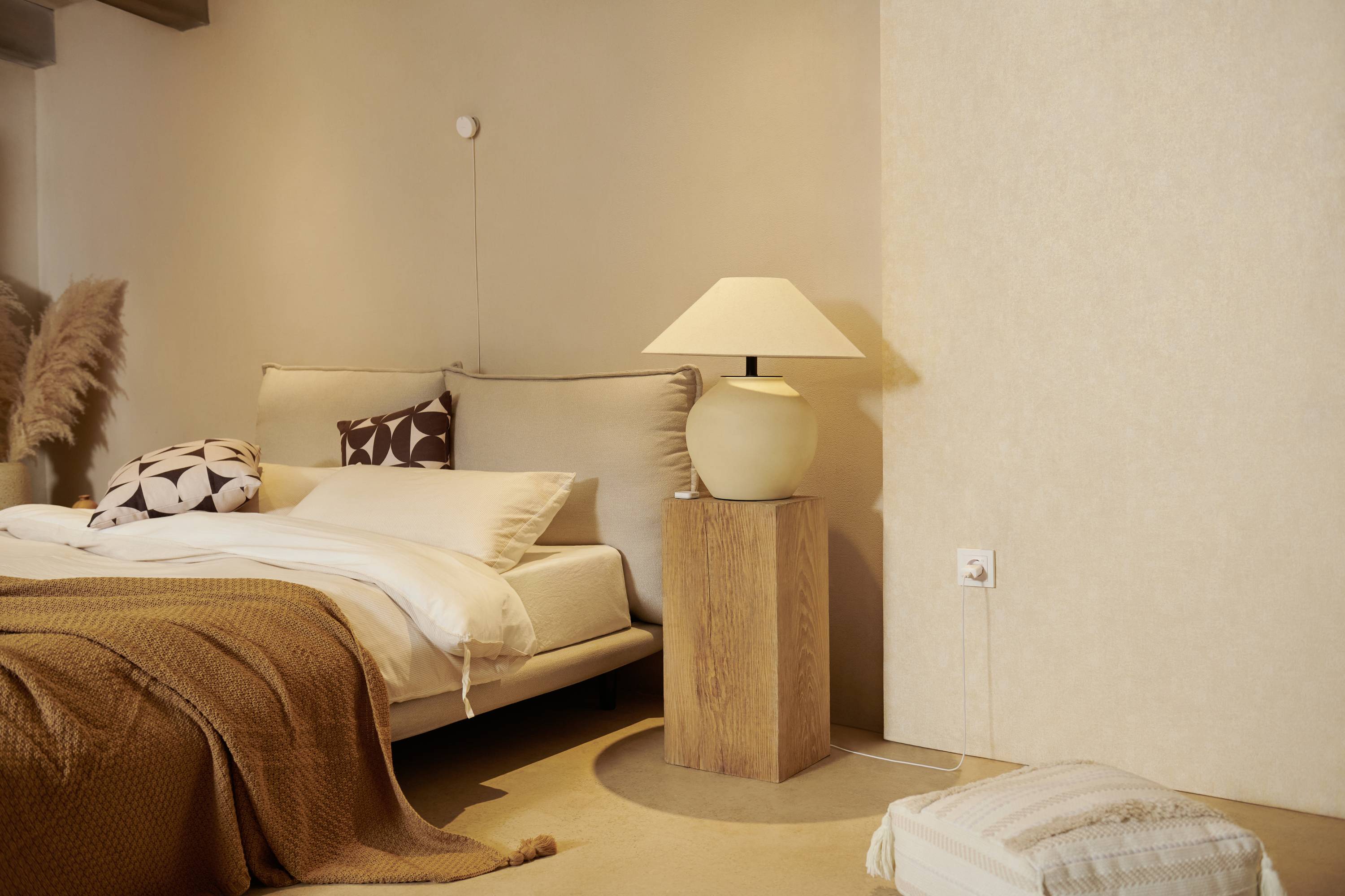 Une chambre confortable avec un lit double, des murs beiges, une lampe de chevet sur une table en bois, et des coussins décoratifs.