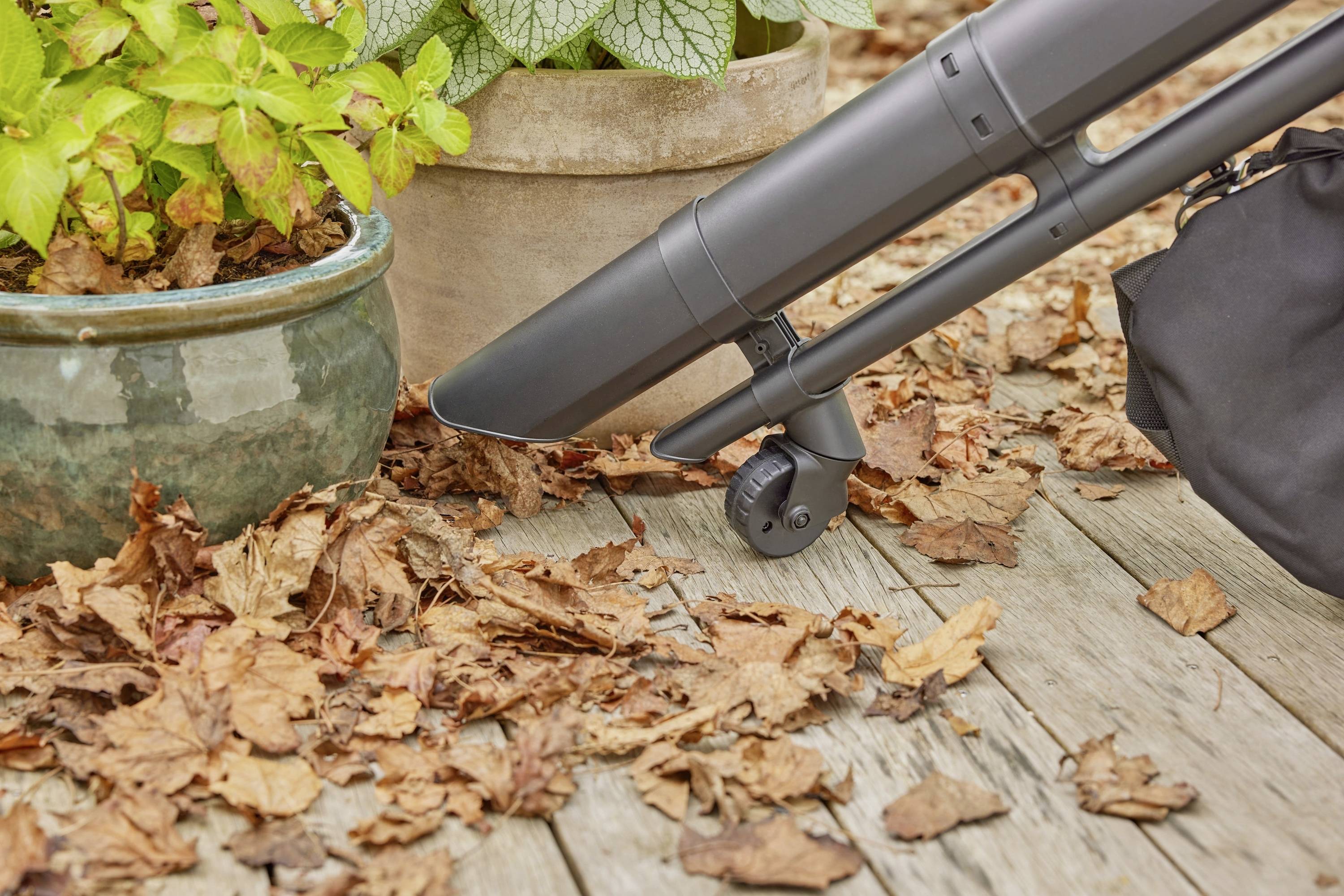 Un souffleur de feuilles enlève des feuilles mortes d'une terrasse en bois. Des plantes en pots sont disposées à côté des feuilles.