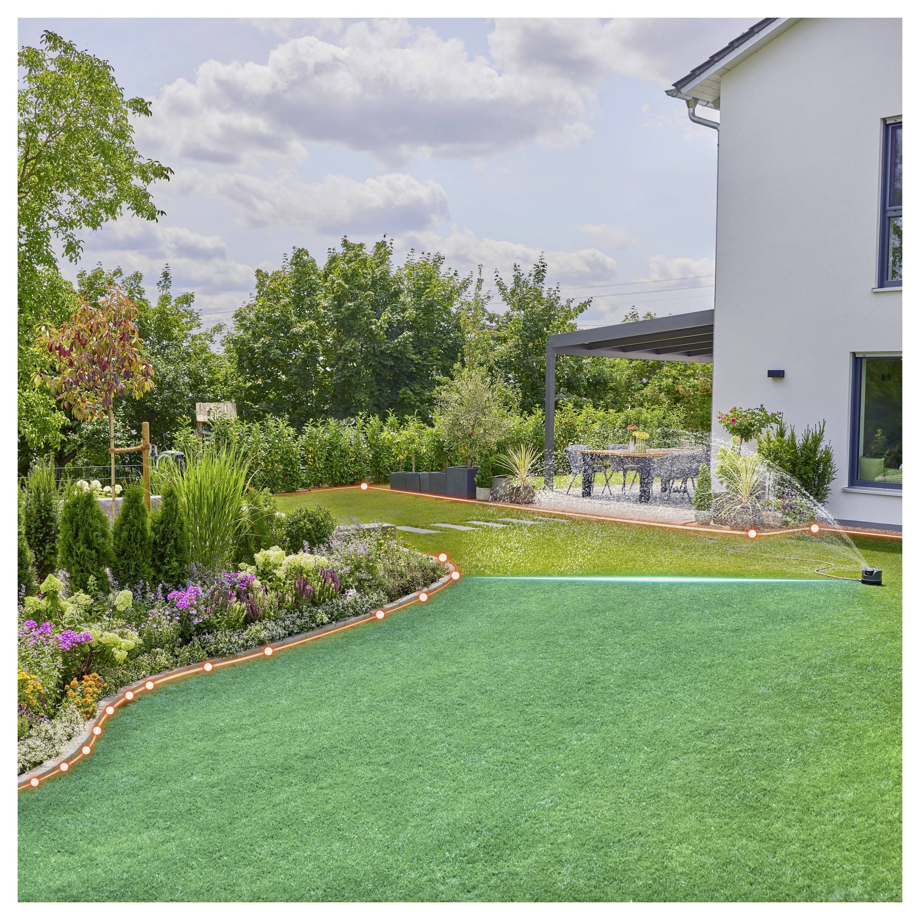 Un jardin luxuriant transforme cette cour arrière avec une pelouse dotée de plates-bandes de fleurs colorées et une zone de patio. Un arroseur irrigue le gazon sous un ciel partiellement nuageux.