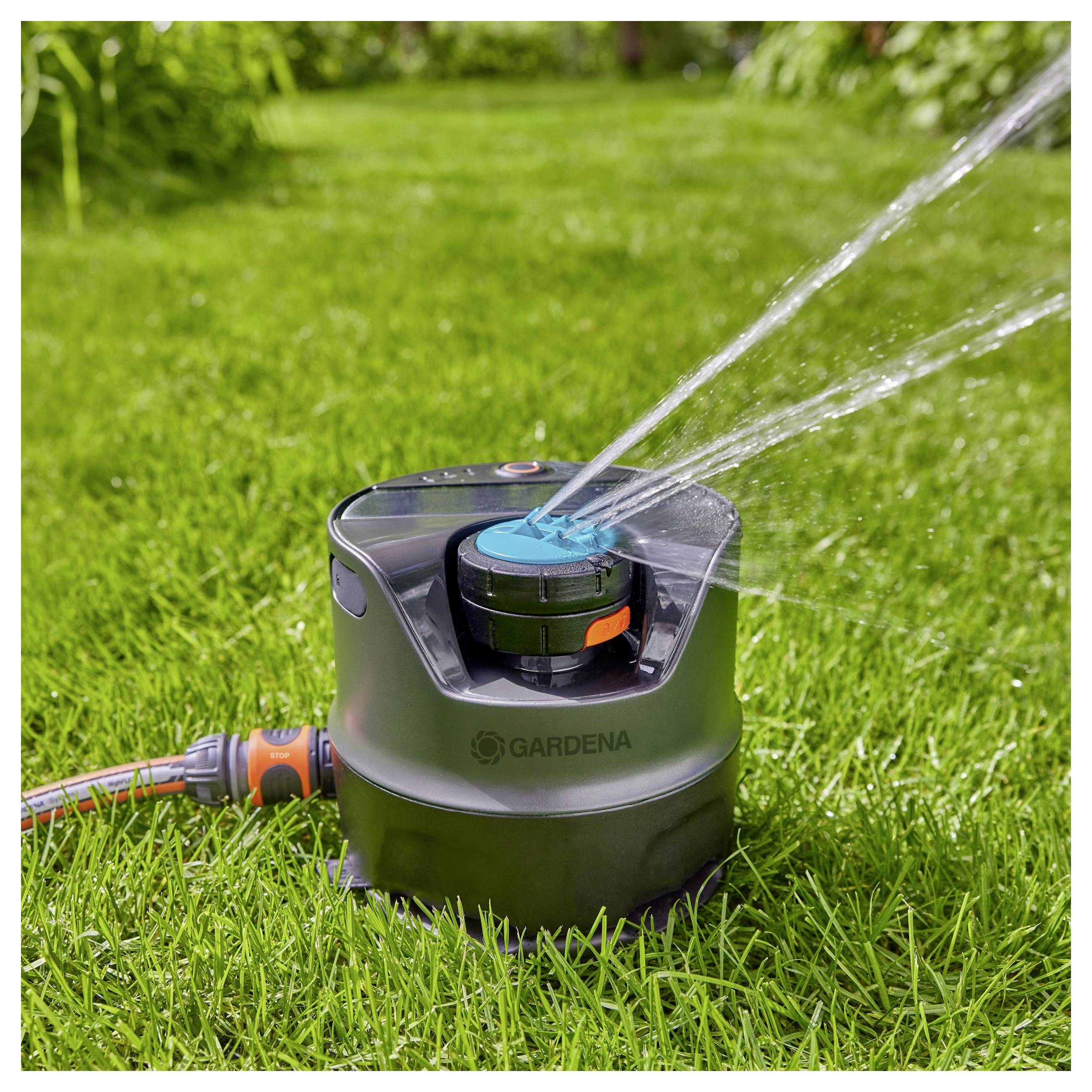 A sprinkler watering grass in a garden, showing water jets spraying in multiple directions. Lush greenery surrounds the sprinkler.