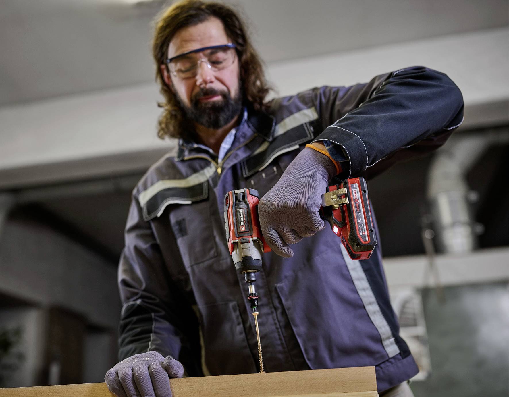 Un homme portant des lunettes de protection et des gants perce un trou dans un morceau de bois à l'aide d'une perceuse électrique dans un atelier.