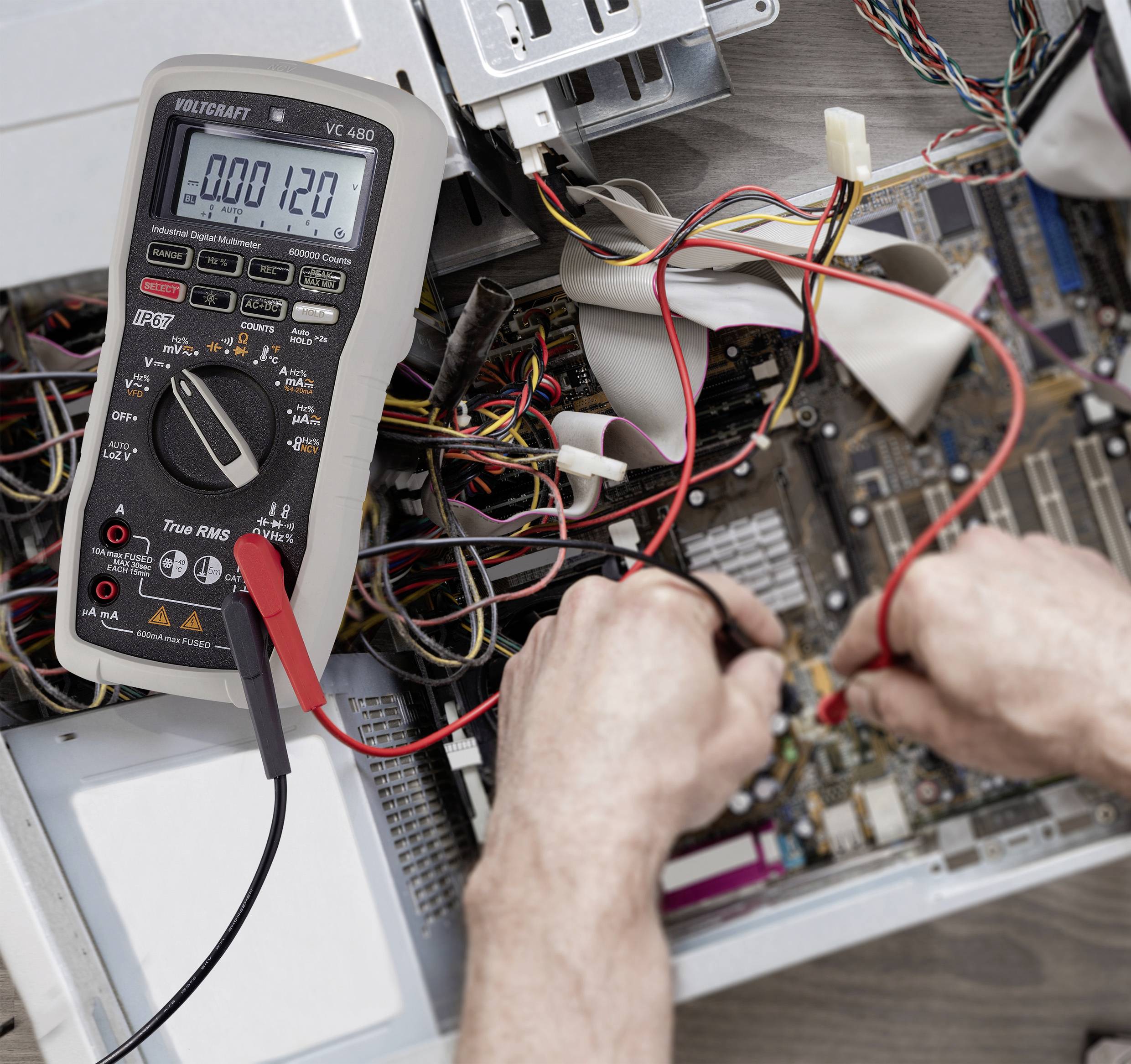 Une personne mesure le courant électrique sur un circuit imprimé ouvert à l'aide d'un multimètre. De nombreux câbles sont visibles.