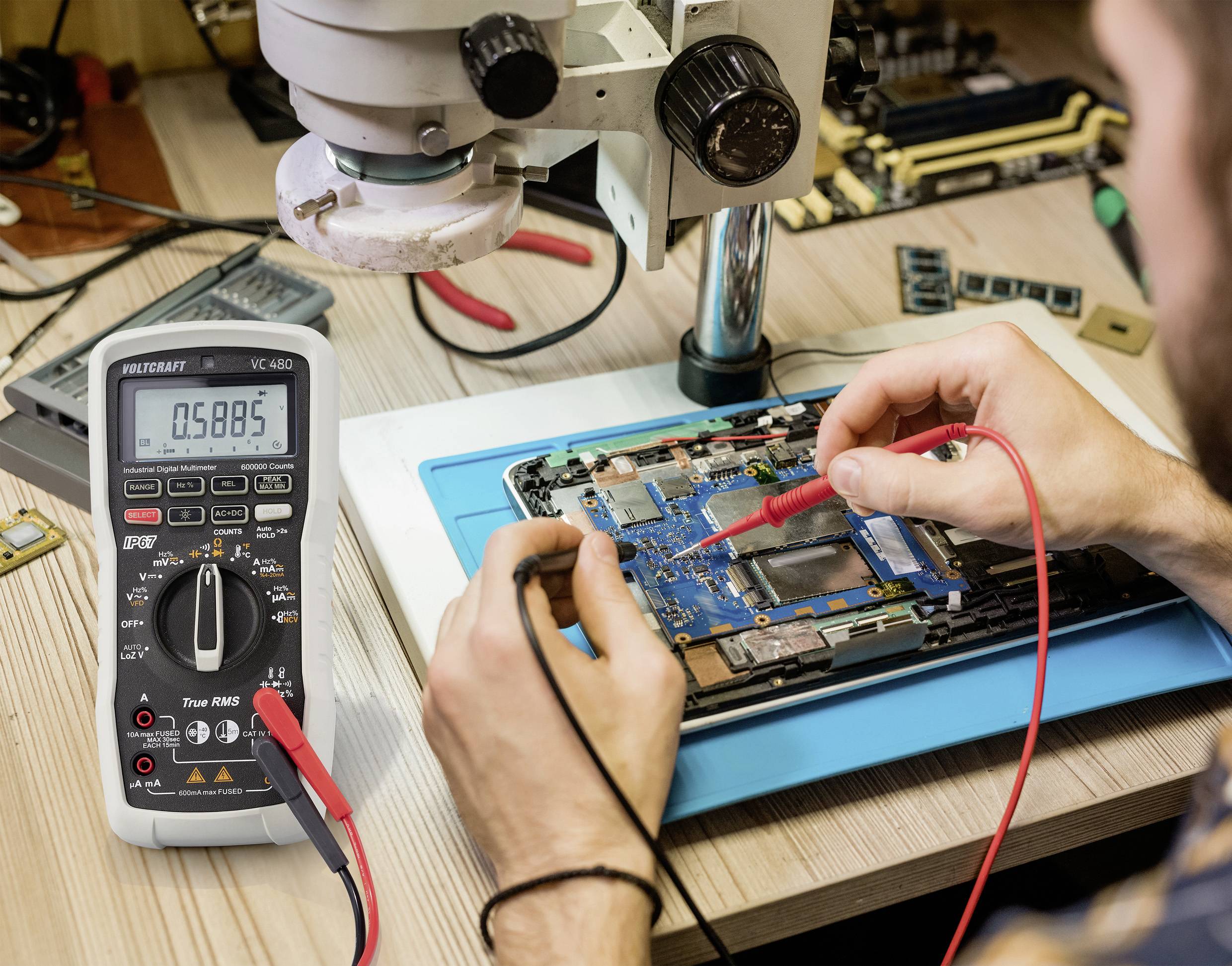Une personne répare une carte électronique sous un microscope, utilisant un multimètre pour mesurer la tension.