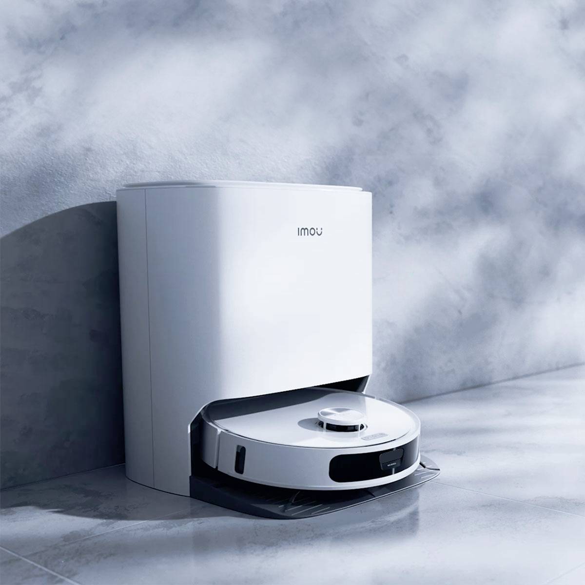 Un aspirateur robot blanc avec station de charge contre un mur, éclairé par la lumière du soleil tombant sur le sol depuis la gauche.