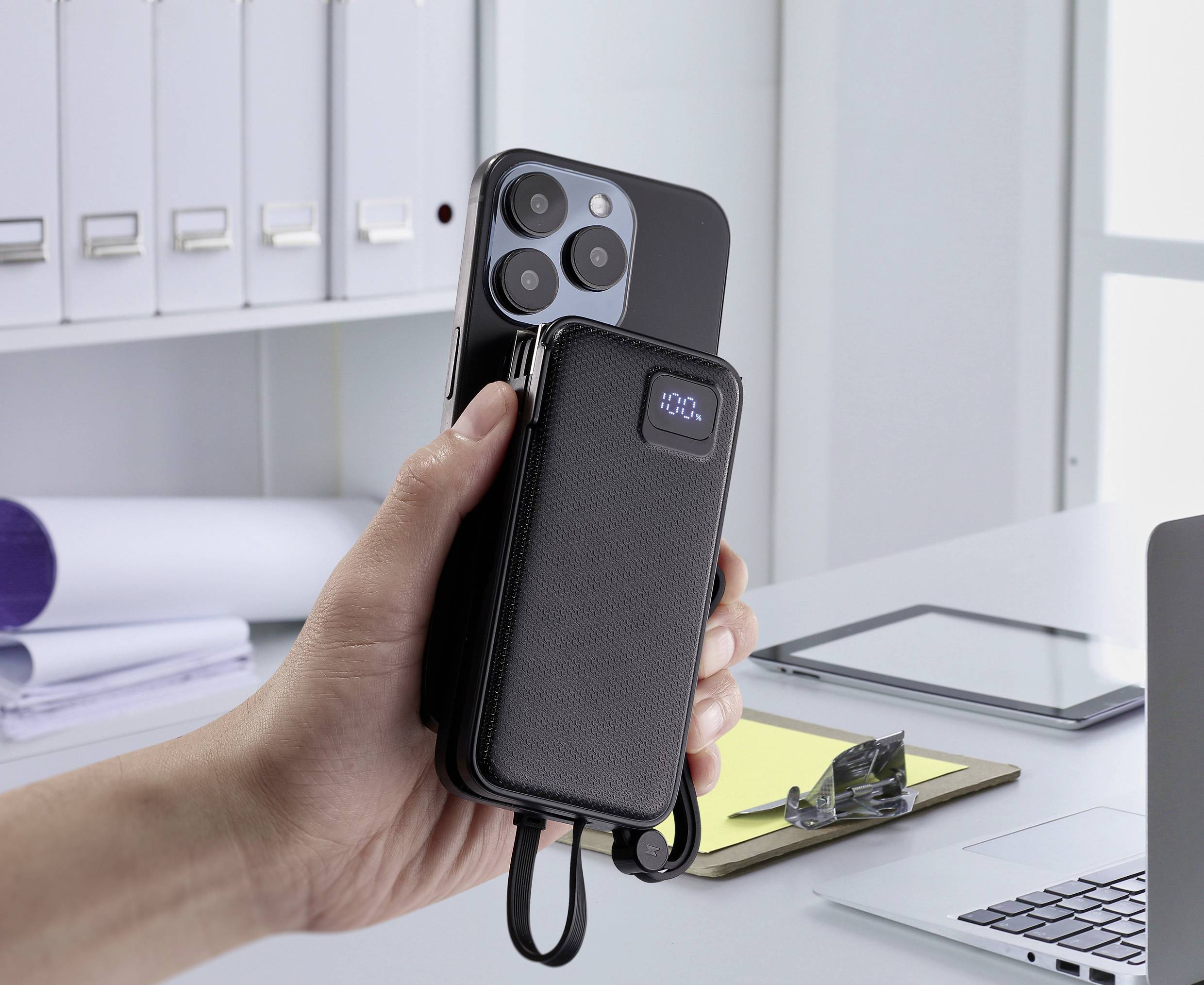 Une main tient un smartphone équipé d'une petite batterie externe connectée, dont l'indicateur affiche une charge complète à 100%. En arrière-plan, un bureau.