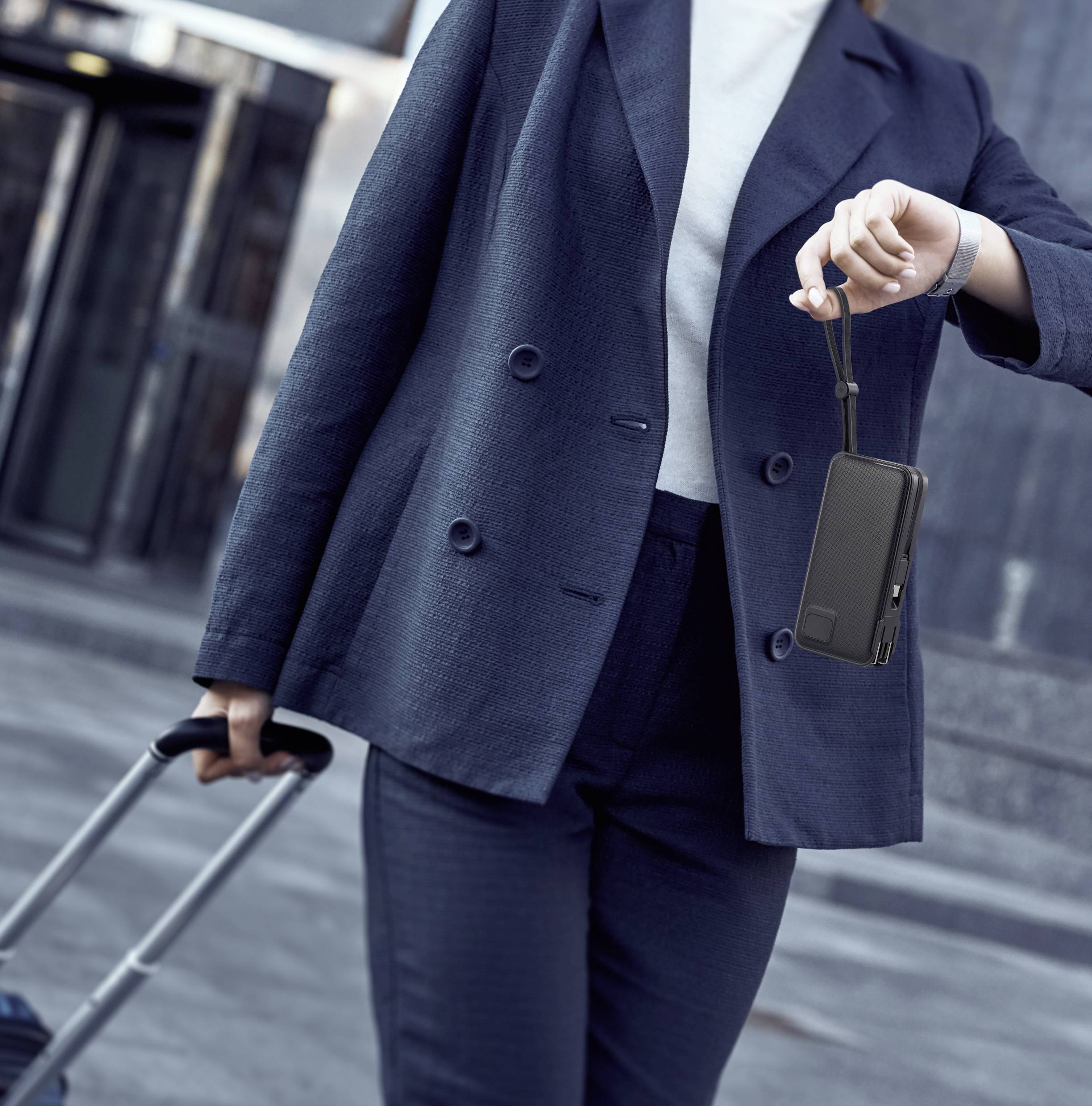 Une personne en costume sombre tire une valise et tient une batterie externe noire à la main. Arrière-plan urbain.