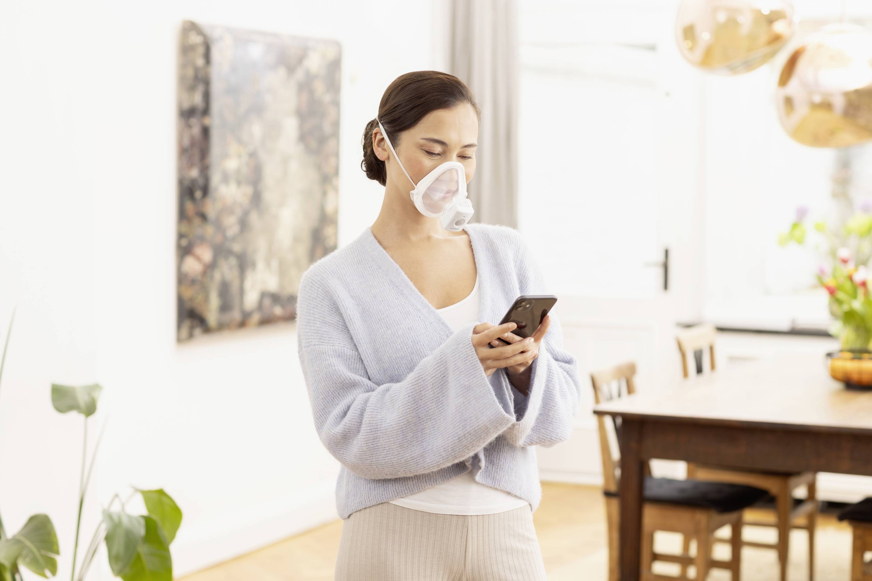 Une personne se tient dans une pièce, portant un masque transparent et regardant un téléphone portable. La pièce est meublée avec des meubles modernes et des œuvres d'art accrochées aux murs.