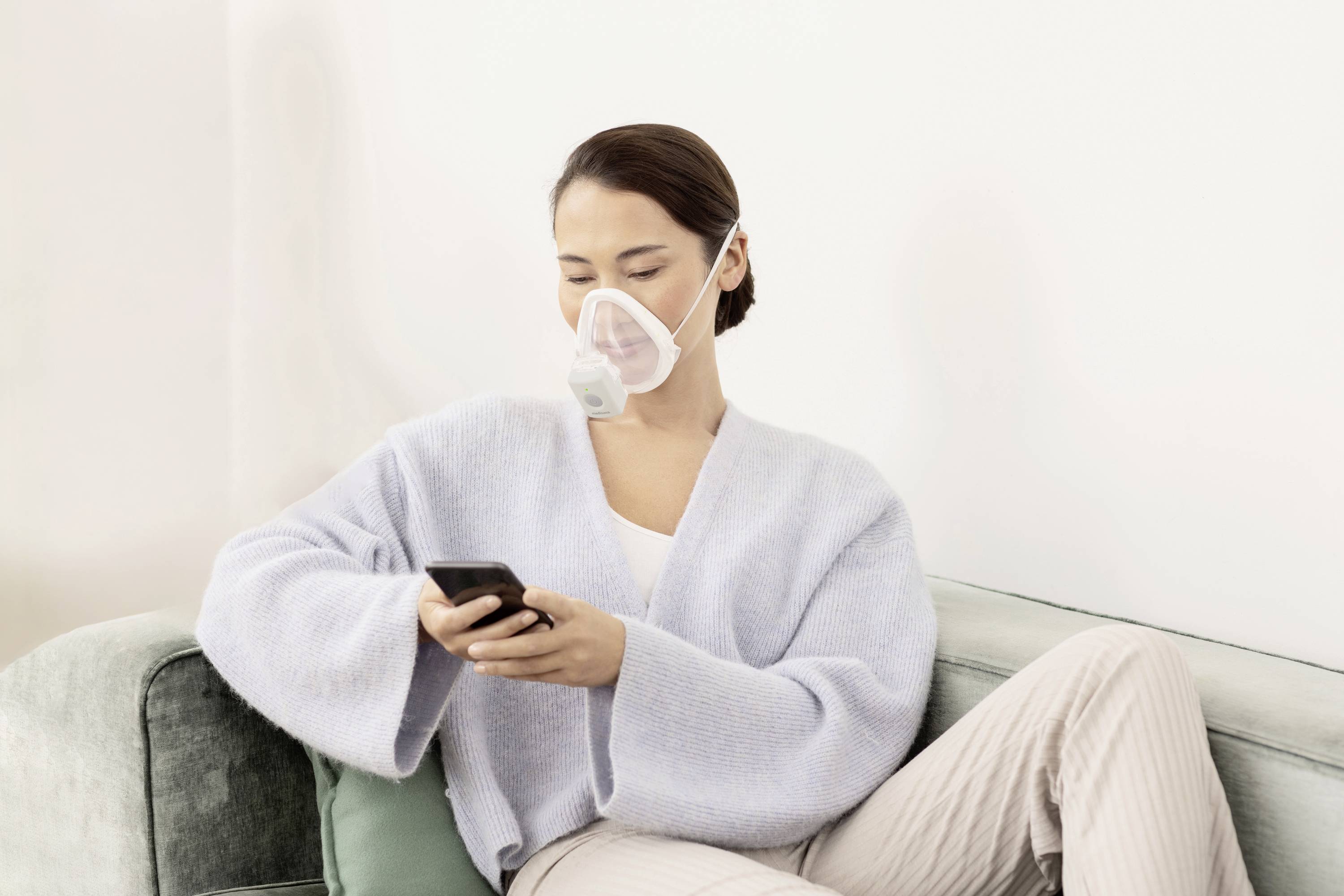 Une personne est assise sur un canapé, porte un masque de protection et utilise un smartphone.