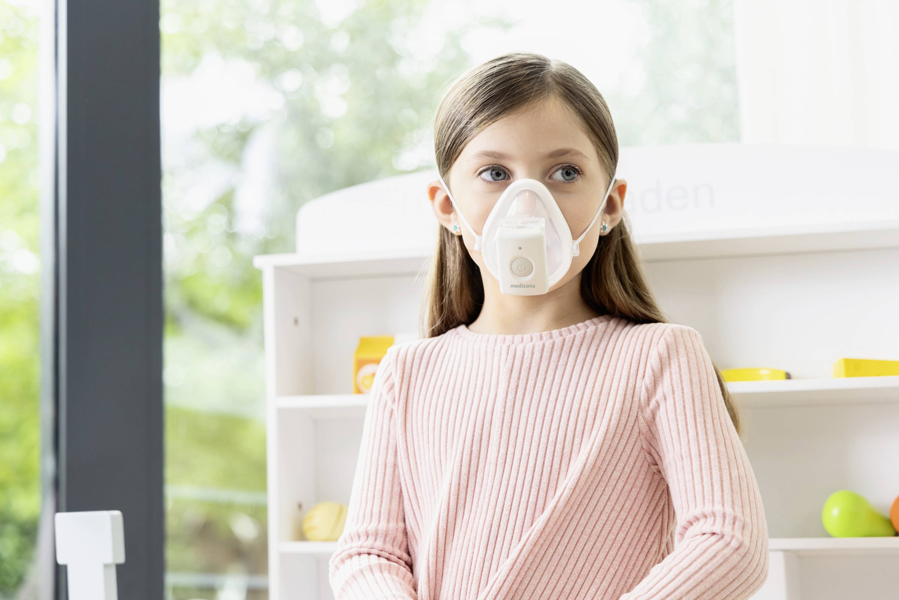 Une fille porte un masque de protection respiratoire dans une pièce claire ; des étagères avec des jouets sont visibles en arrière-plan.