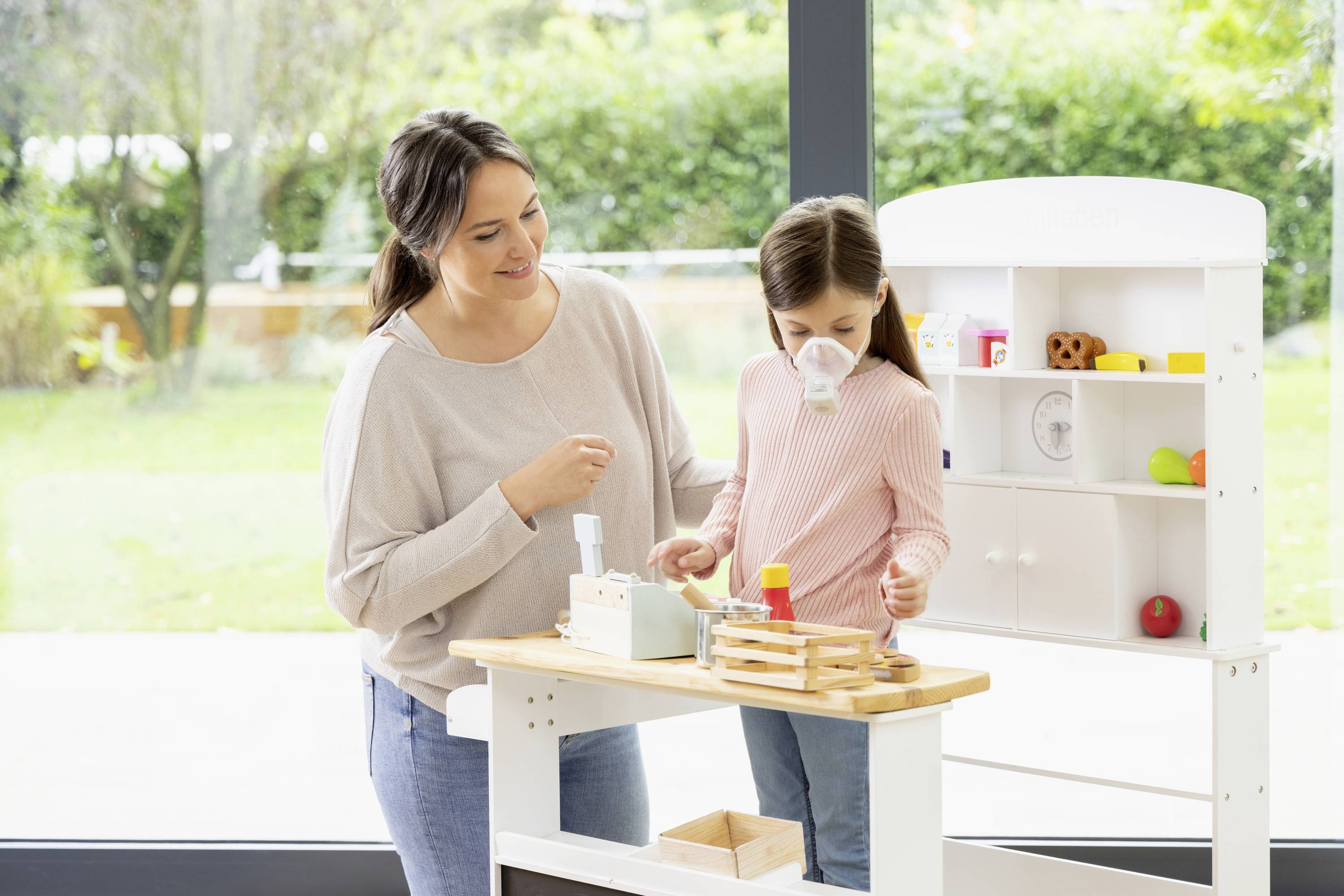 Une femme et un enfant jouent avec une cuisine pour enfants devant une grande fenêtre. L'enfant porte un masque de déguisement.