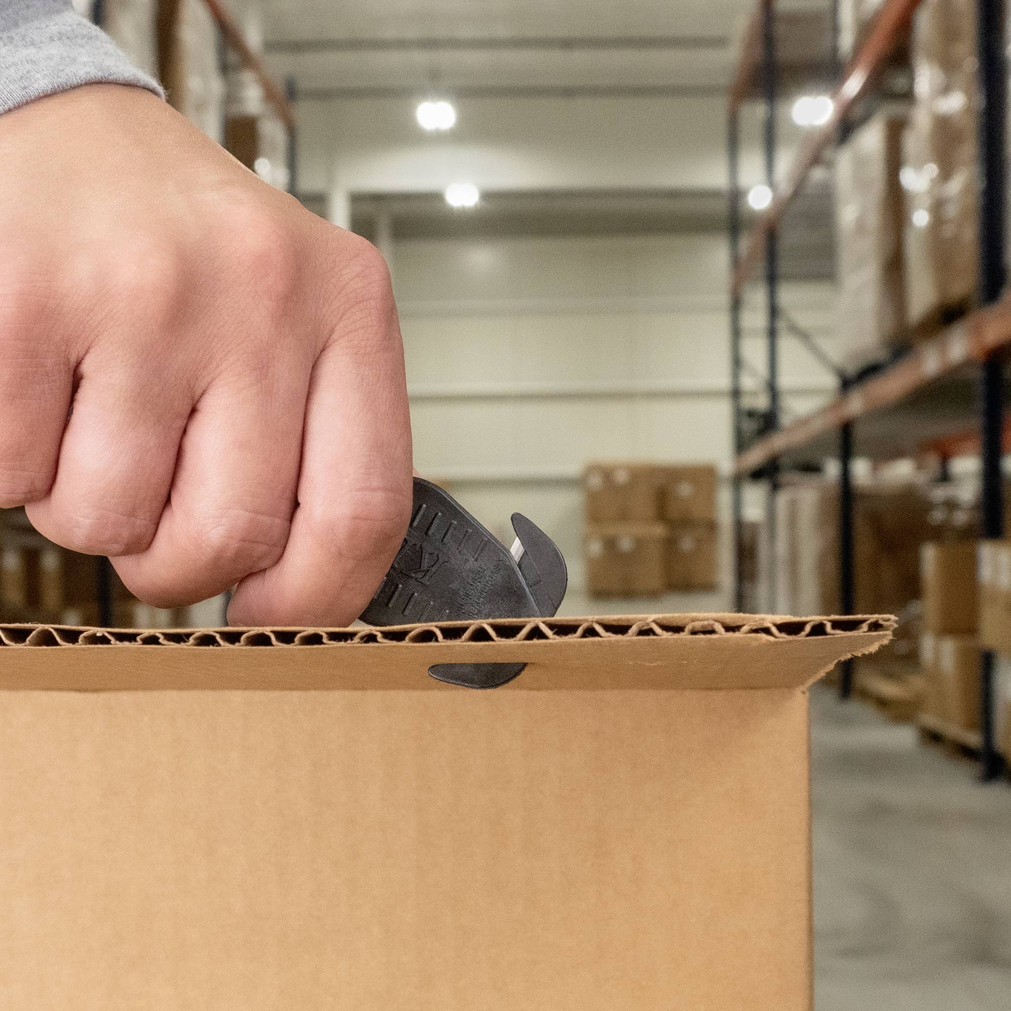 Une personne ouvre un carton avec un cutter dans un entrepôt. À l'arrière-plan, on aperçoit des étagères chargées d'autres cartons.