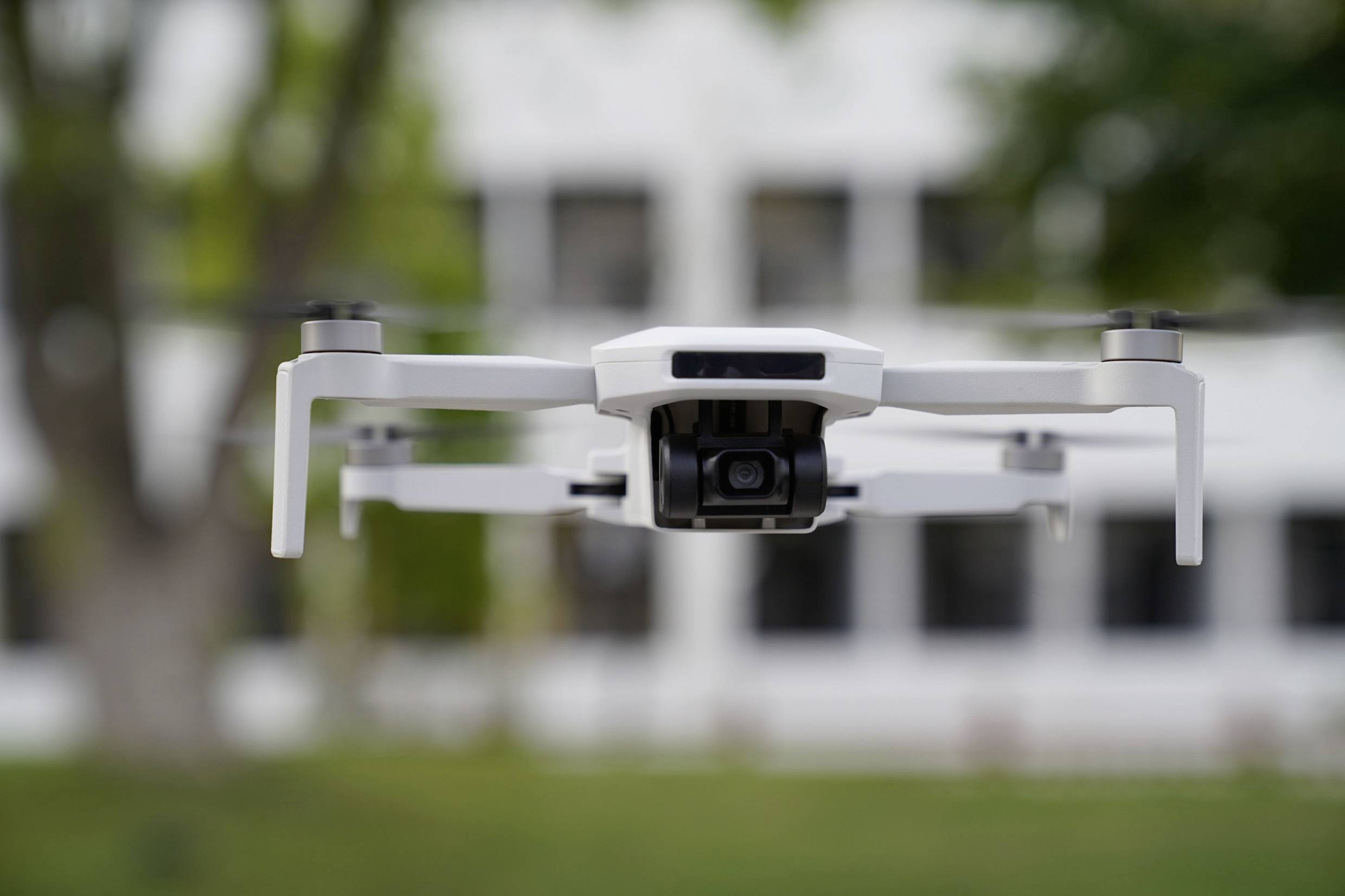 Un drone blanc plane dans les airs devant un arrière-plan flou composé d'arbres et d'un bâtiment à plusieurs étages.