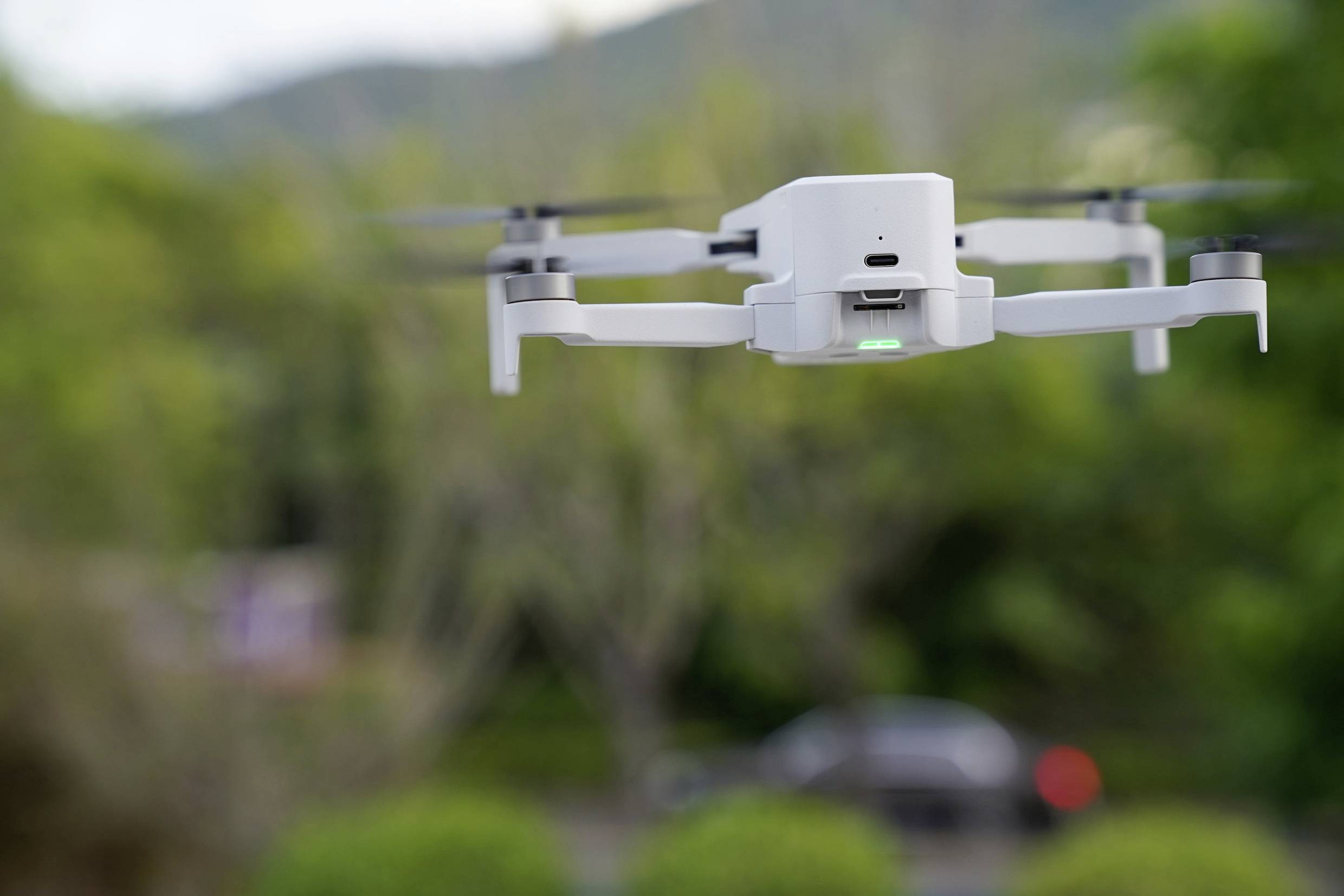 Un drone blanc plane à l'extérieur devant un paysage vert flou. Il a quatre rotors et une lumière verte brillante à l'avant.