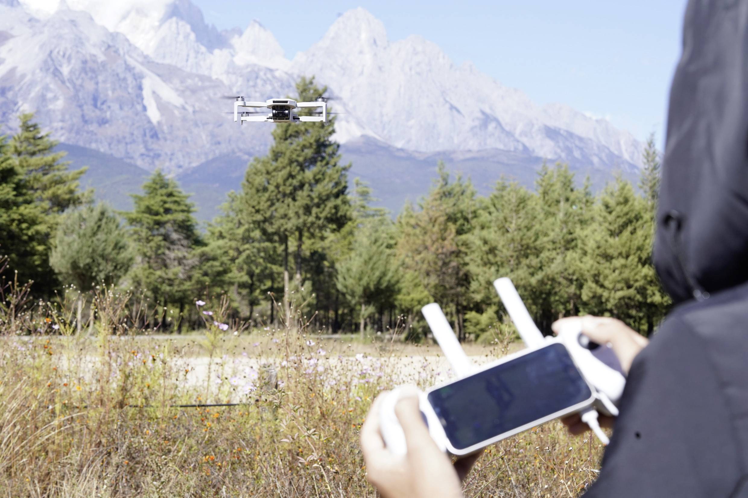 Une personne pilote un drone à l'extérieur à l'aide d'une télécommande. Le drone plane devant un paysage montagneux boisé.
