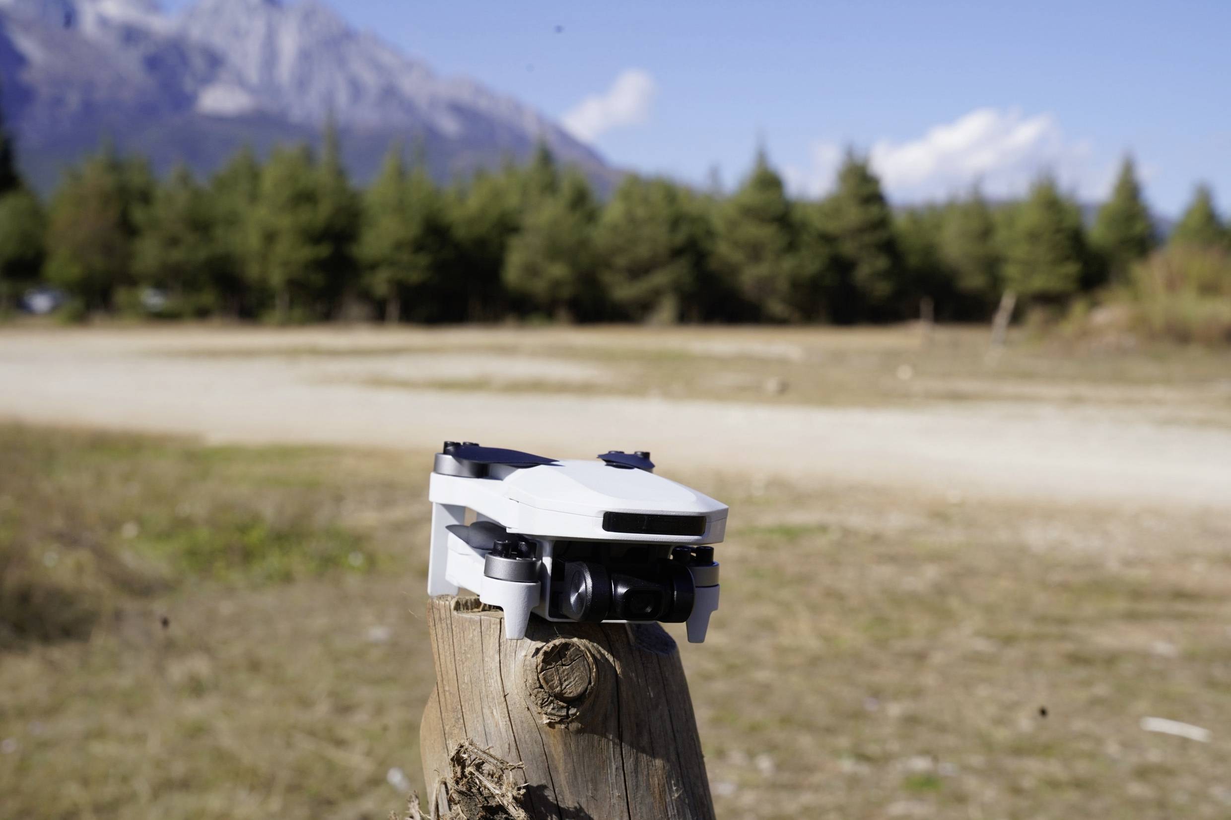 Un petit drone blanc est posé sur un poteau en bois dans un paysage forestier avec des montagnes en arrière-plan sous un ciel bleu.