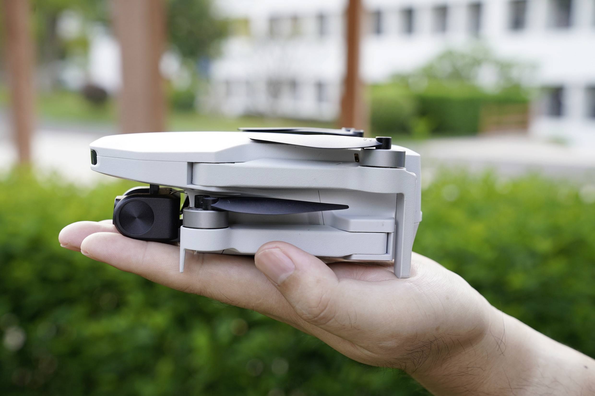 Une main tient un petit drone plié à l'extérieur. En arrière-plan, des arbres et des bâtiments flous sont visibles.