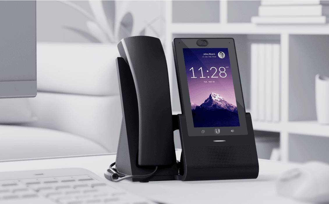 Un téléphone de bureau noir moderne avec un grand écran affiche l'heure 11:28 et l'image d'une montagne. Un bureau se trouve en arrière-plan.