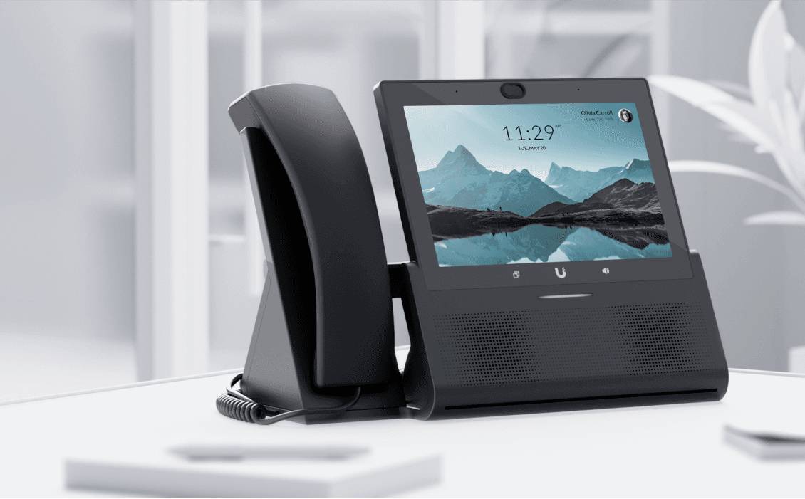 Téléphone noir à écran tactile affichant 11h29 et un paysage montagneux. Placé sur une table blanche dans un bureau moderne.