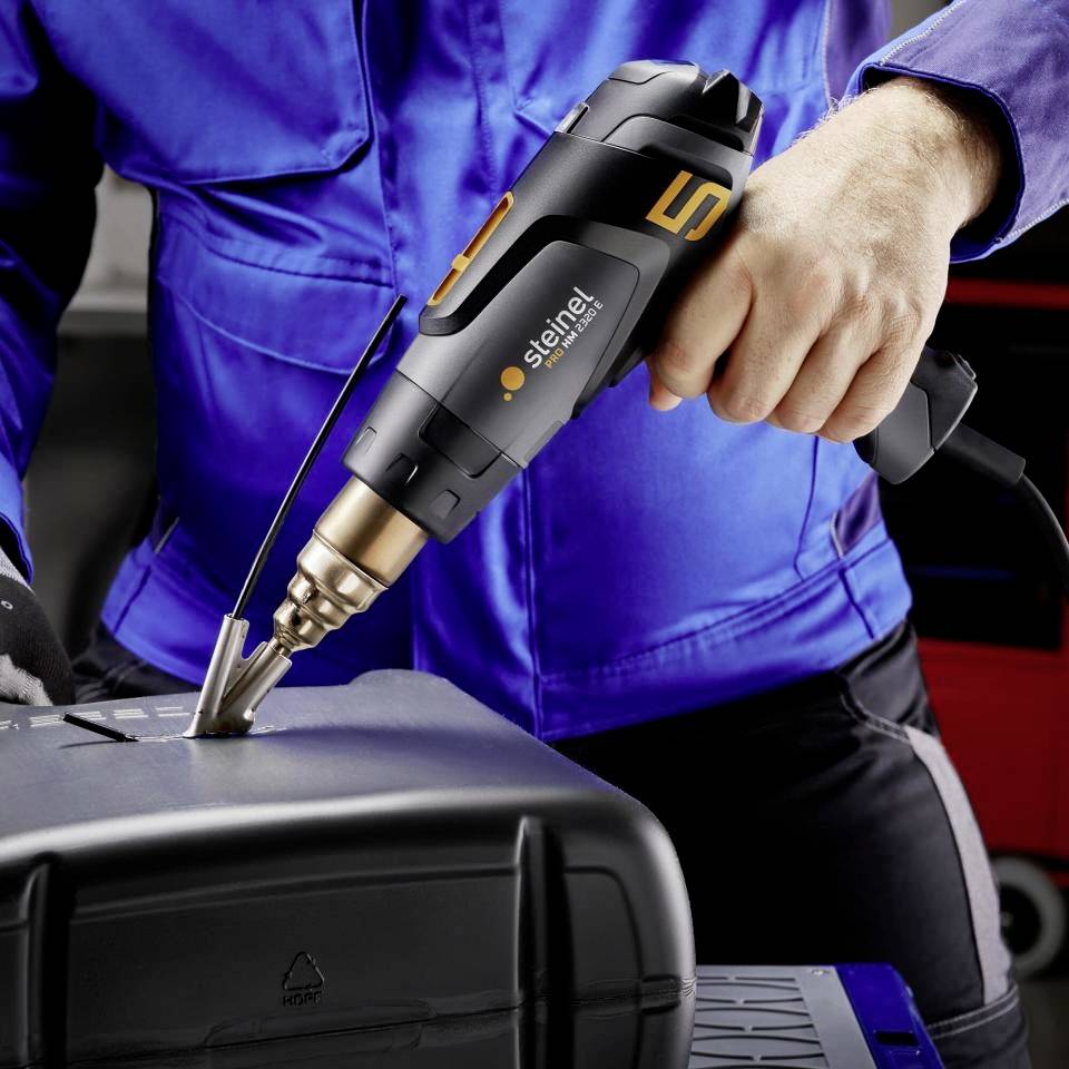 Un ouvrier en tenue de travail bleue utilise un pistolet à air chaud noir pour fondre ou traiter un matériau.