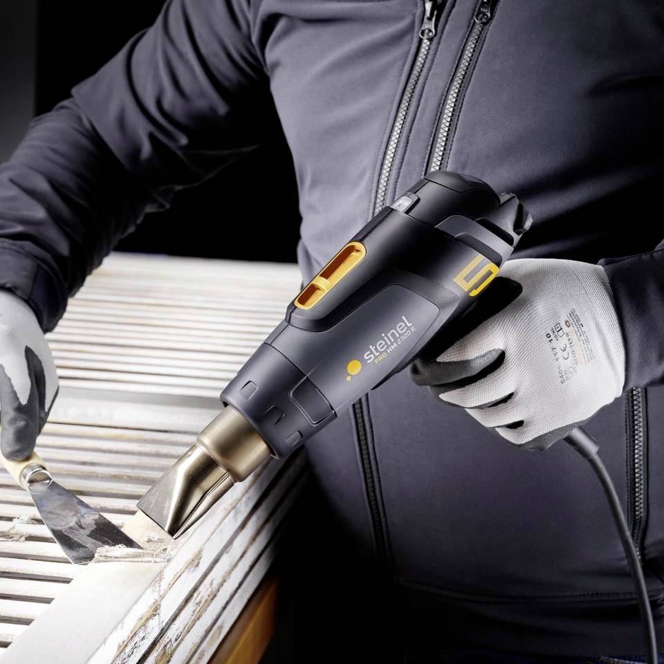 Une personne utilise un pistolet à air chaud sur une surface en bois, portant des gants. L'outil de marque 'Steinel' est destiné à des travaux de rénovation ou de réparation.