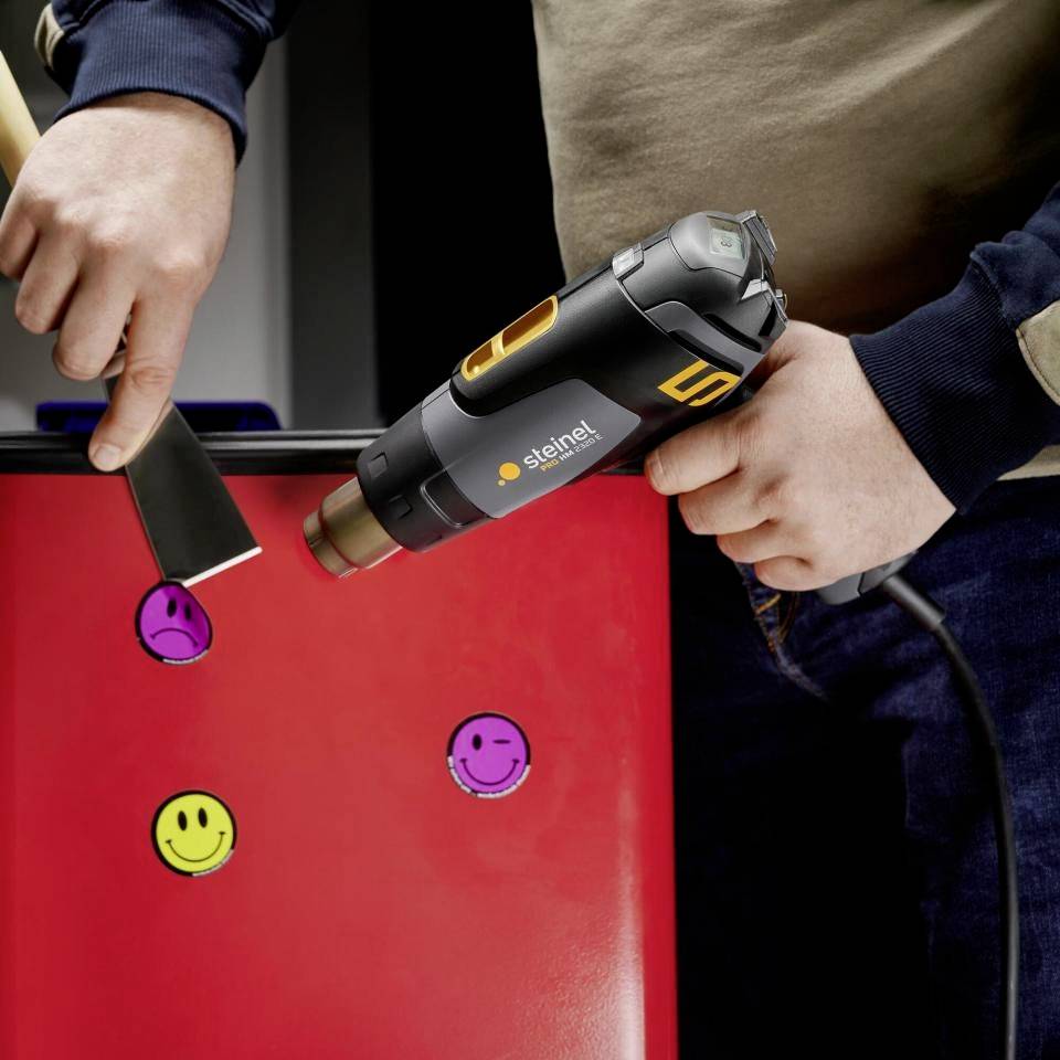 Une personne tient un sèche-cheveux dirigé vers un objet rouge avec des autocollants colorés représentant des visages.