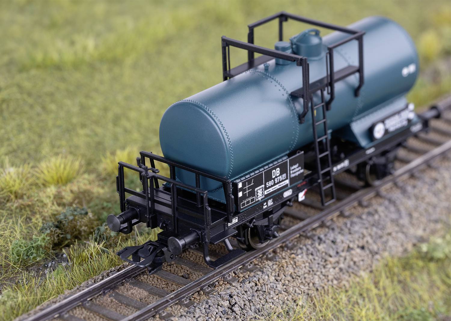 Un modèle d'un wagon-citerne vert sur un rail de chemin de fer miniature, entouré d'herbe et de gravier.