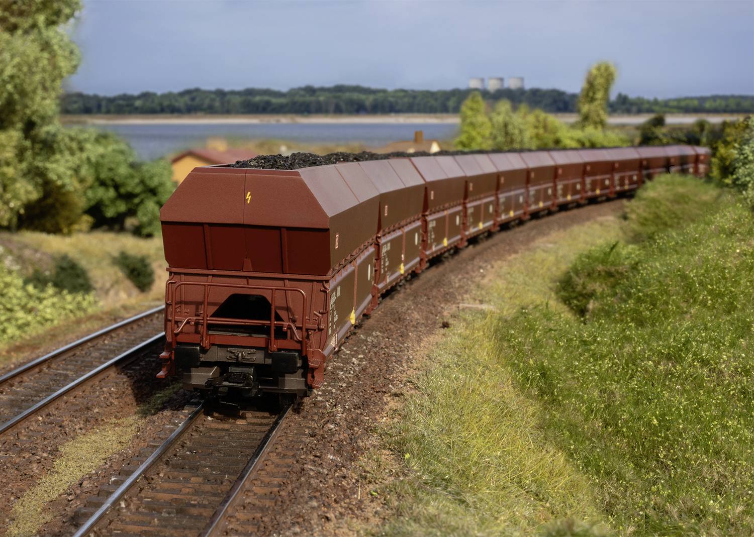 Un long train de marchandises avec des wagons ouverts remplis de charbon circule le long d'une voie ferrée rurale. Des arbres et un lac sont visibles en arrière-plan.