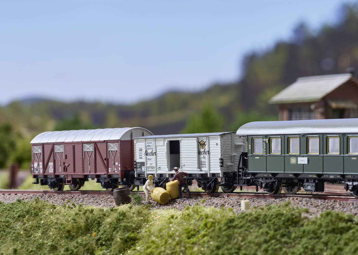 Réseau ferroviaire miniature avec une scène fidèle aux détails : Deux personnages réparent les voies ferrées à côté d'un train composé de trois wagons, dans un environnement paysager.