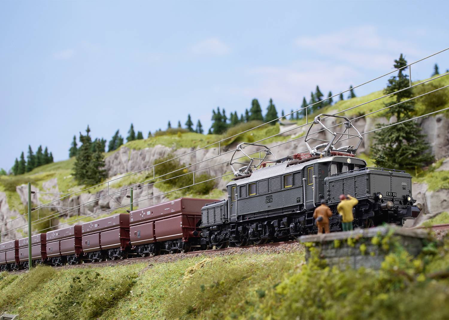 Un train miniature avec un train de marchandises traversant un paysage parsemé d'arbres et de collines. Deux personnages sont debout à côté du train.