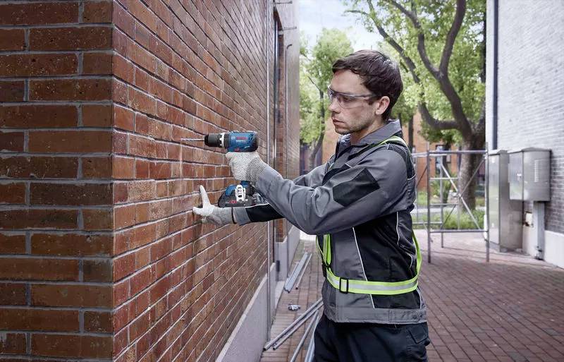 Un homme portant des lunettes de protection et des gants de travail fore un mur de briques à l'extérieur à l'aide d'une perceuse électrique.